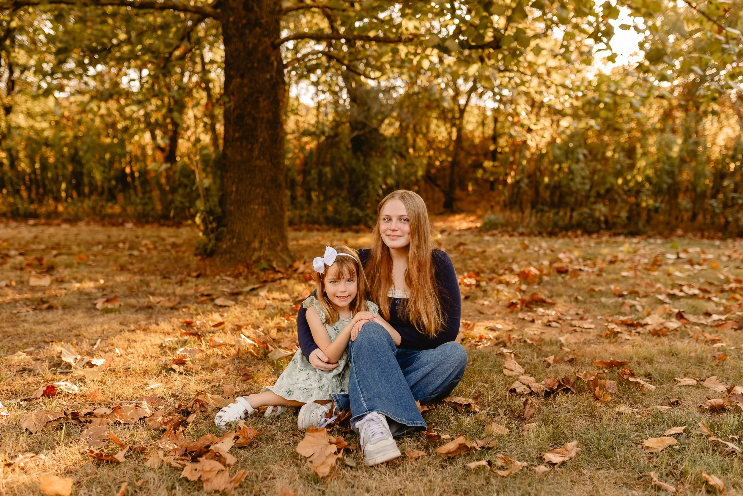 A woman and a young girl sitting on the ground in a park surrounded by fallen autumn leaves. The woman has long reddish hair and is smiling gently at the camera. The girl has long blonde hair tied with a white bow and is smiling widely. There are trees with yellow and orange leaves in the background.