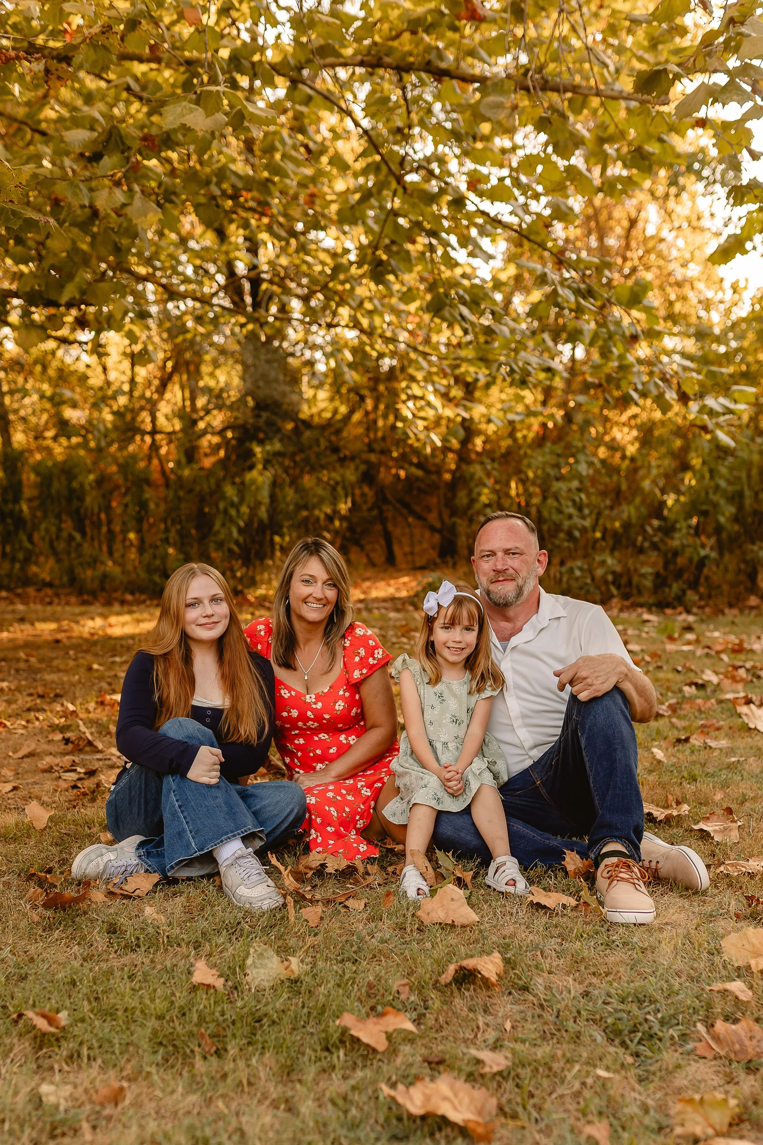 A family of five sitting on the grass in a park with autumn leaves, smiling at the camera.