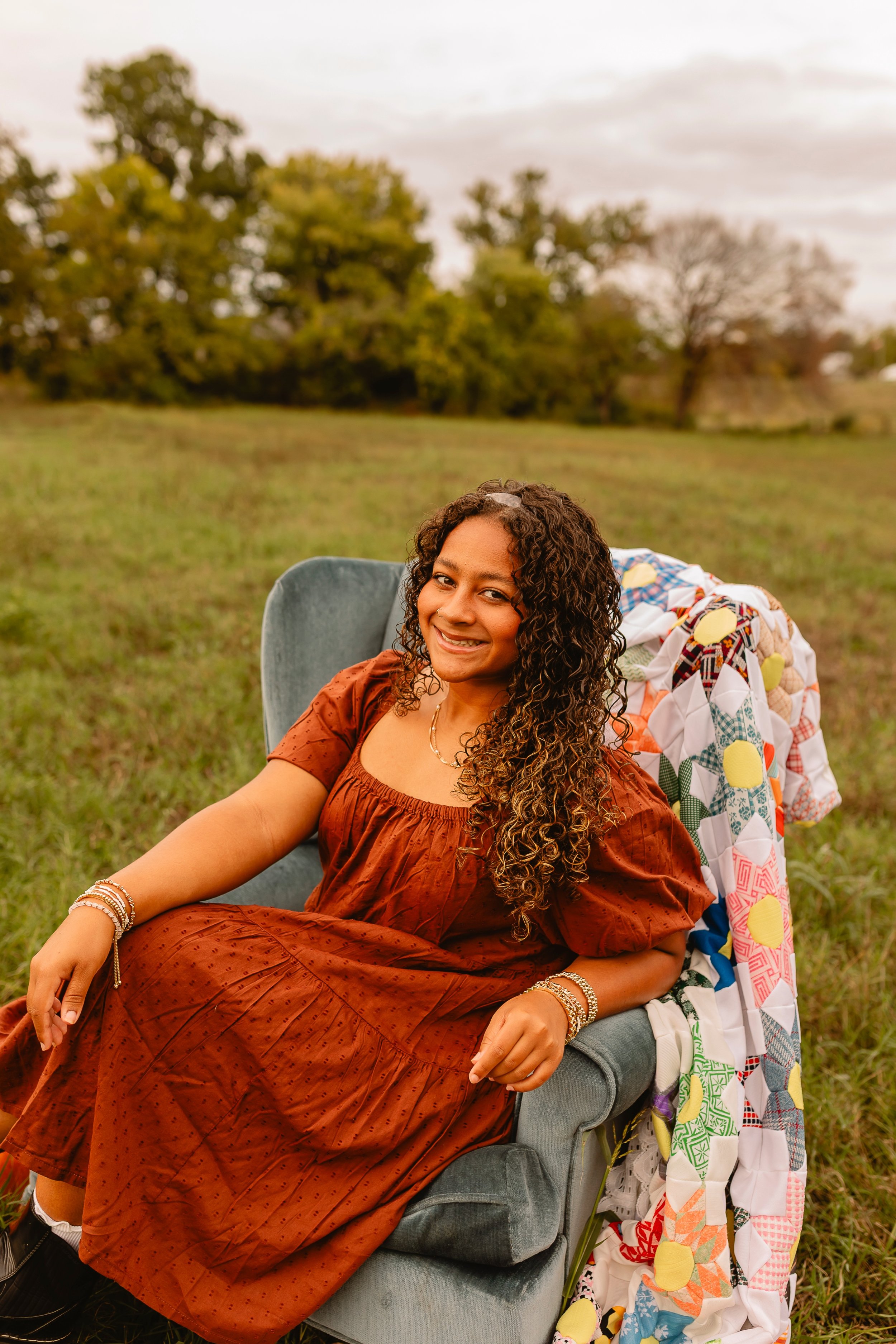 A woman with curly hair, wearing a rust-colored dress, sitting in a gray armchair outdoors in a grassy field during daytime, smiling at the camera. There is a colorful patchwork quilt draped over the back of the chair, with a background of trees and 