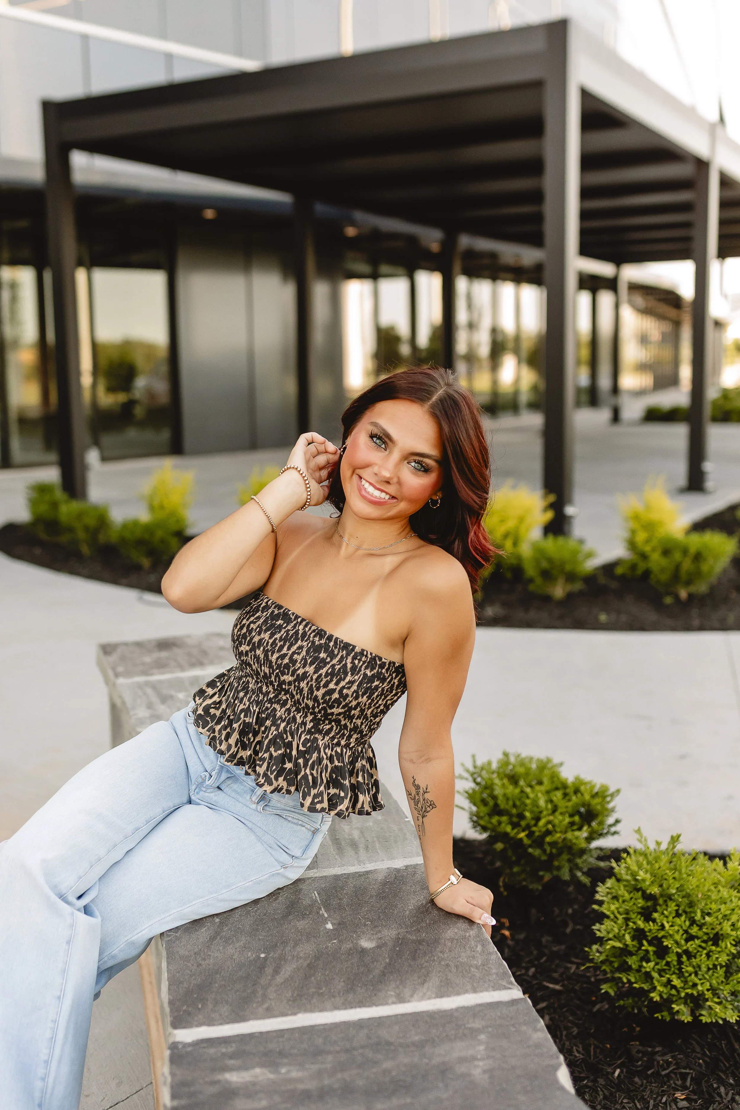 A woman sitting on a stone bench in an outdoor urban setting, smiling at the camera, wearing a leopard print top and light blue jeans, with a modern building and landscaped greenery in the background.