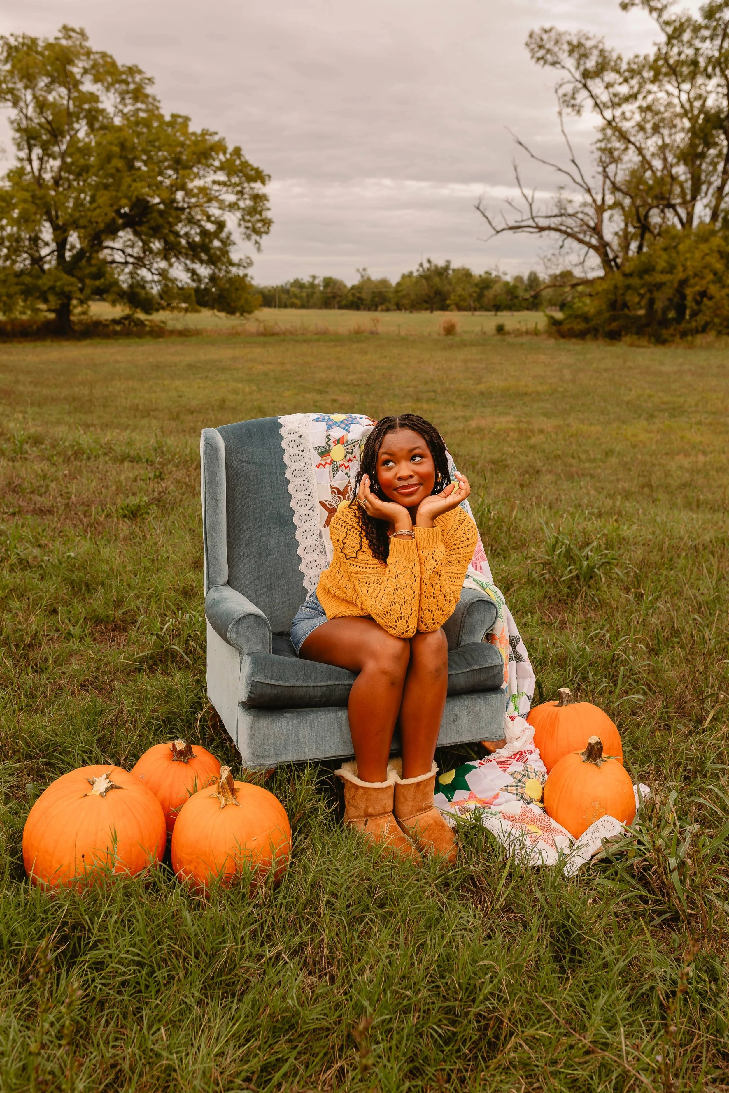 Young woman sitting on a vintage armchair outdoors in a pumpkin patch, surrounded by pumpkins, with trees and cloudy sky in the background.