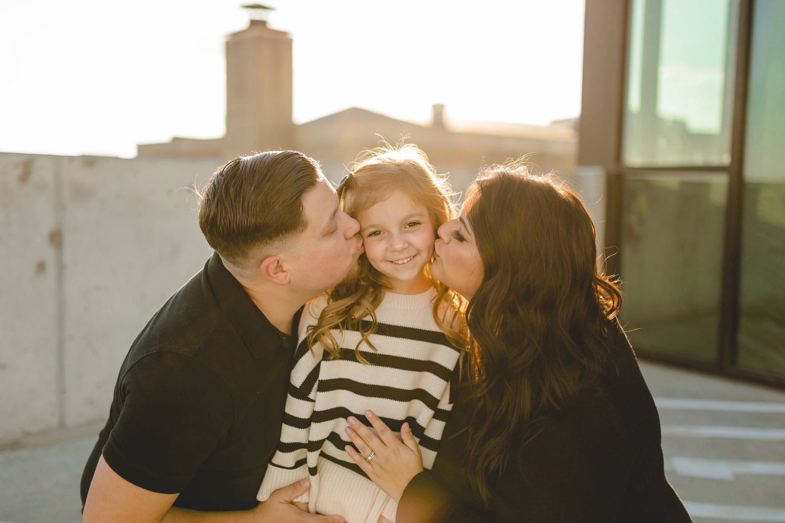 Three people, a man, a woman, and a young girl, sharing a moment of affection outdoors on a sunny day.