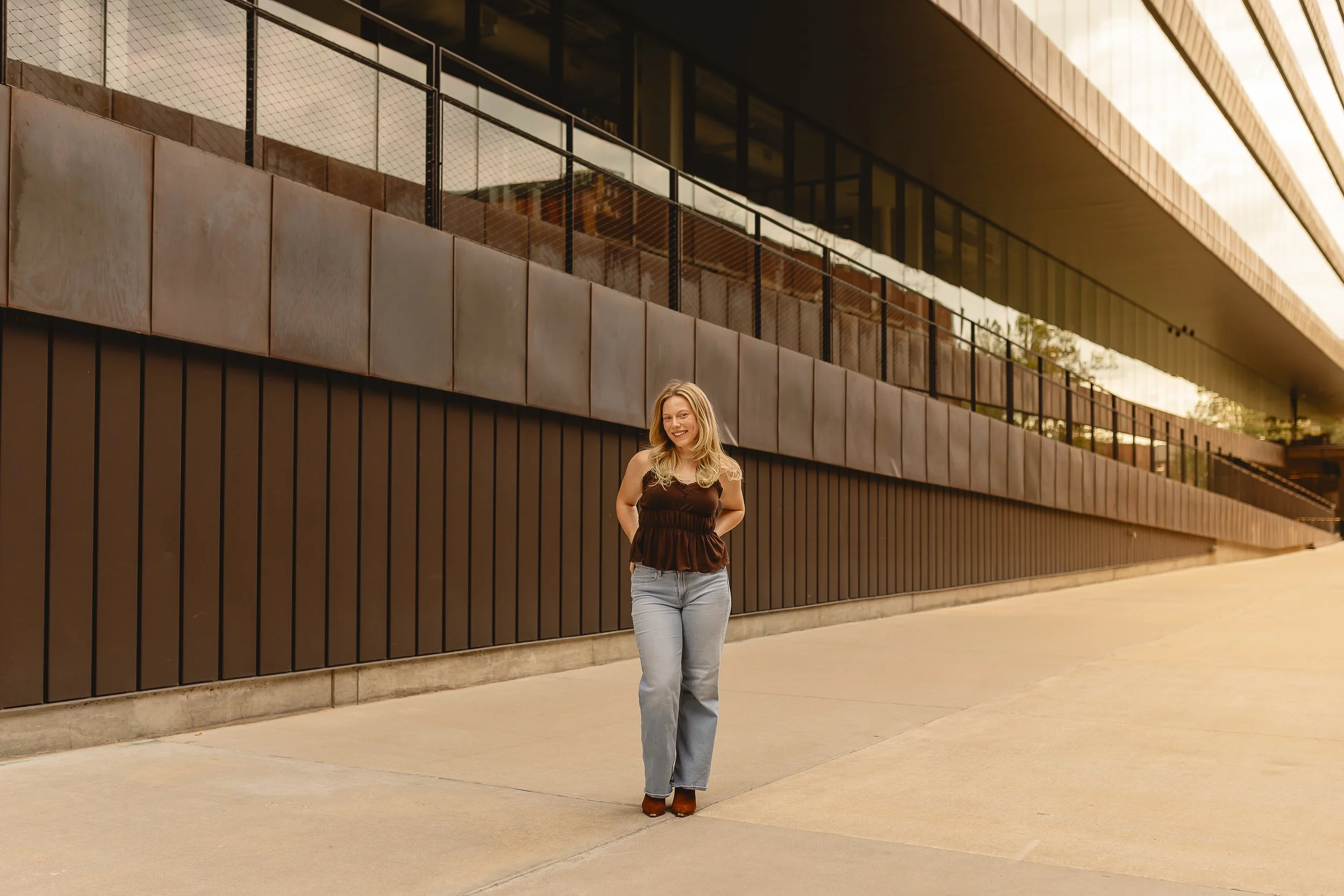 A woman smiling and walking along a modern building with large glass windows and black metal accents, wearing a black sleeveless top and light blue jeans.