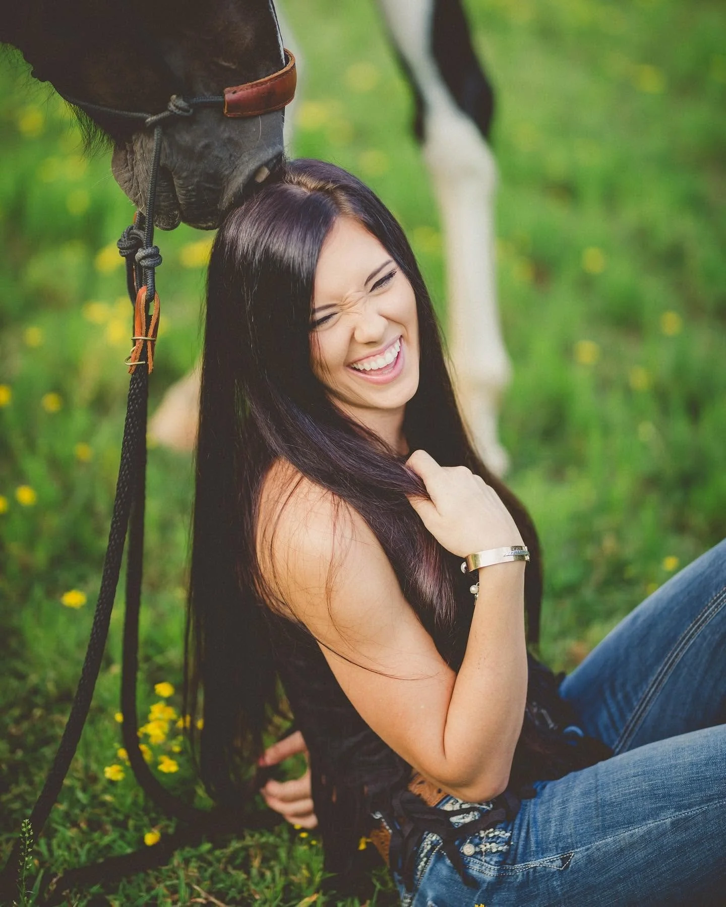 Young woman with long dark hair smiling and trading a kiss with a horse in a grassy field with yellow flowers. The woman is wearing a black tank top, blue jeans, and a silver bracelet.