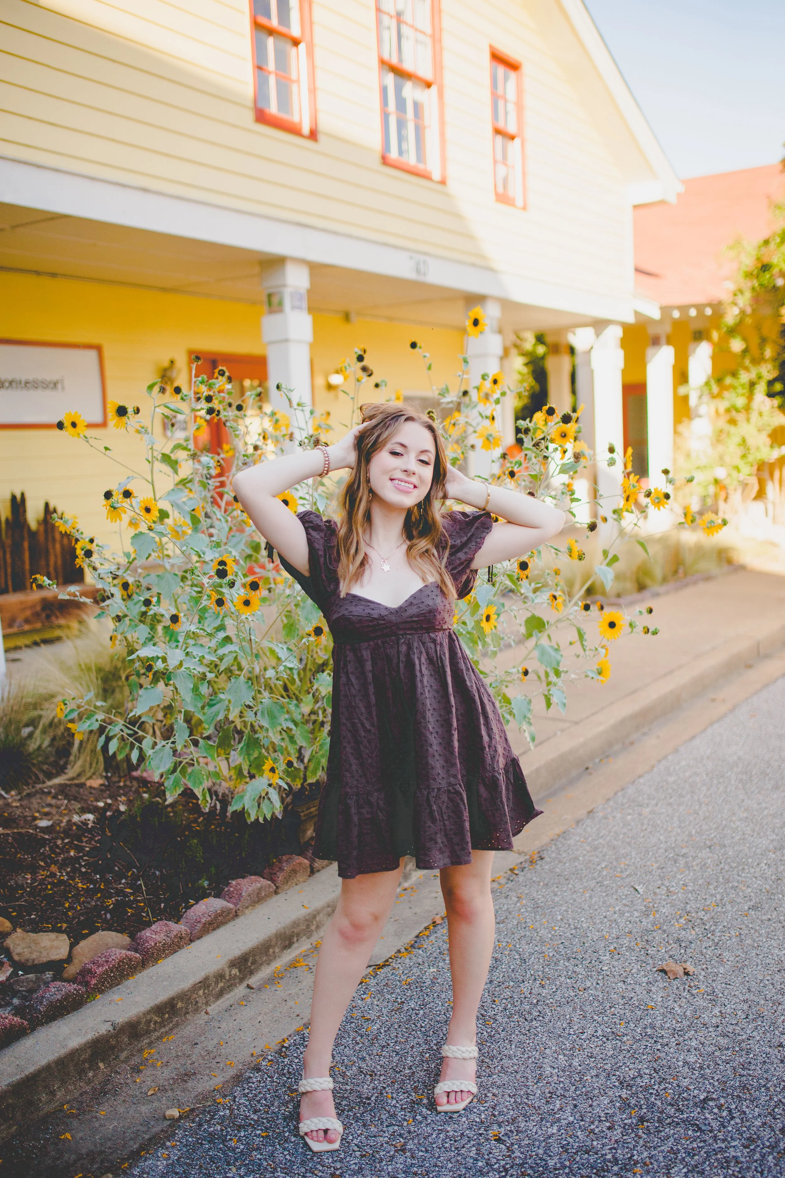 A woman standing on a sidewalk in front of a yellow house with red-trimmed windows and sunflower plants. She is smiling, with her hands behind her head, wearing a dark purple dress and white high-heeled sandals.