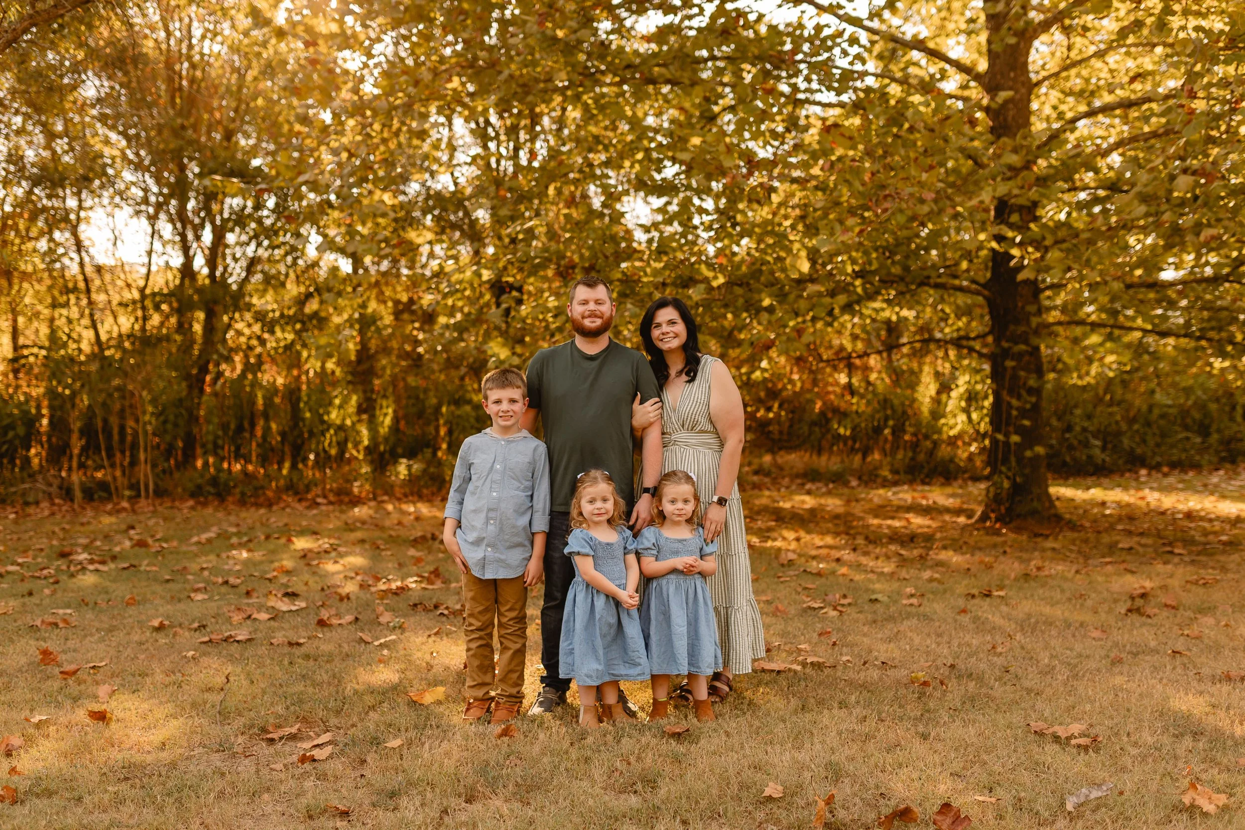 A family of seven standing outdoors in a park during autumn, with fall leaves on the ground and trees with yellow and brown leaves in the background, all smiling.