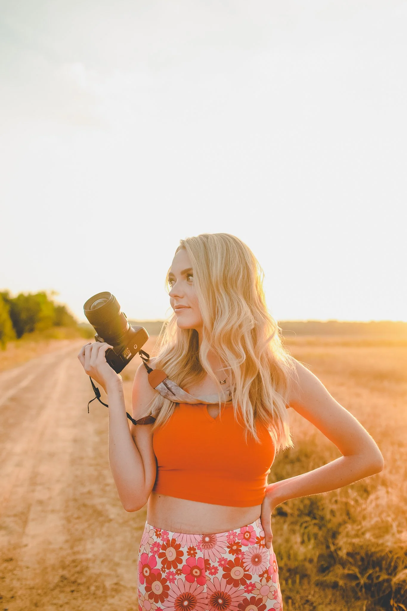 A woman with long blonde hair wearing an orange crop top, floral skirt, and holding a camera, standing in a rural field during sunset.