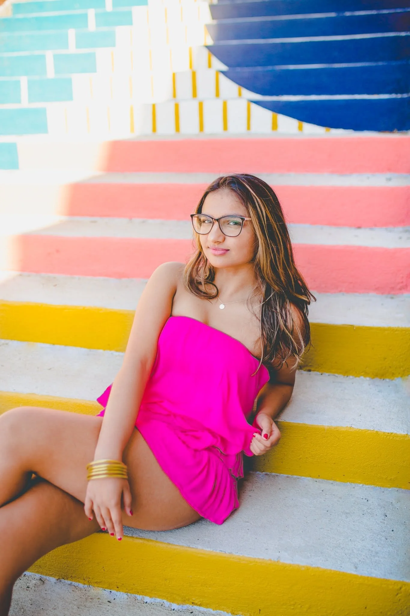 A young woman in a pink dress with glasses sitting on colorful stairs outdoors.