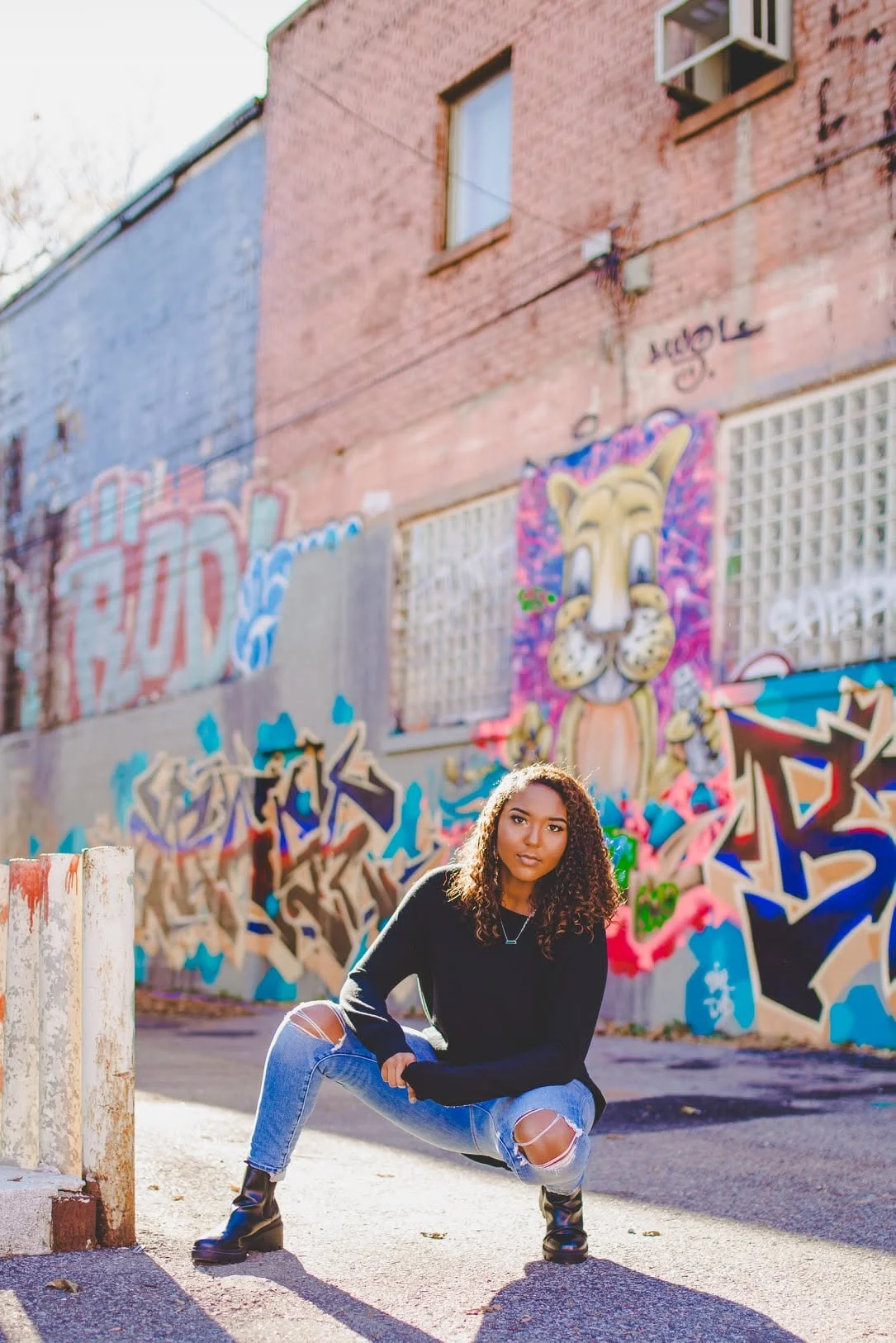 Young woman with curly hair crouching in front of a colorful graffiti mural on an urban wall, wearing black sweater, ripped jeans, and black boots.