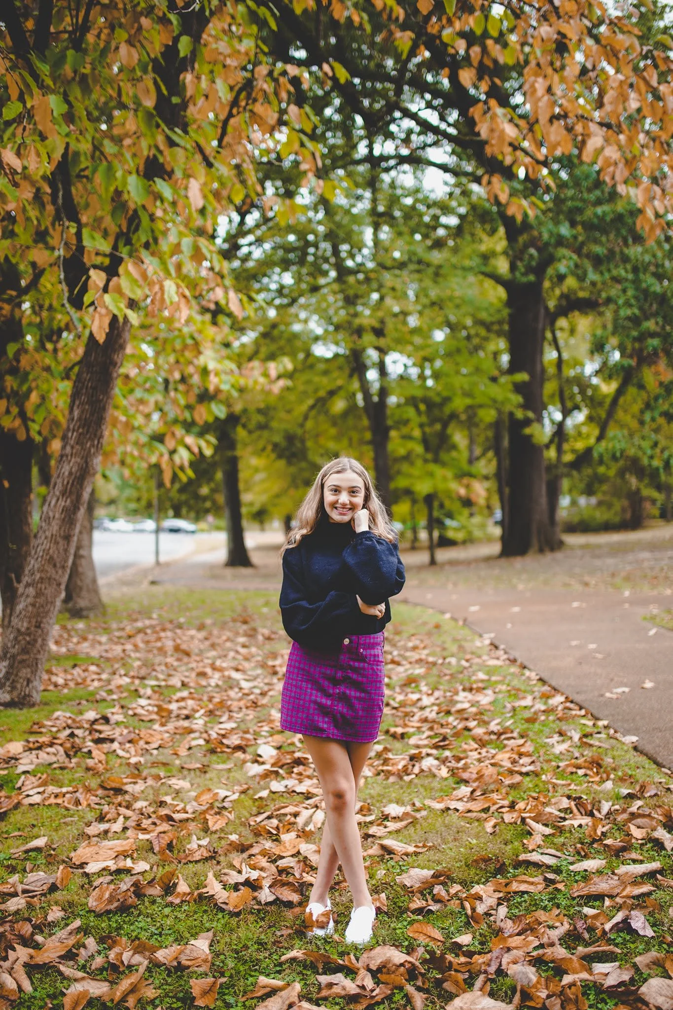 A young woman with long hair smiling and walking in a park during autumn, surrounded by fallen leaves and trees with green and orange leaves.