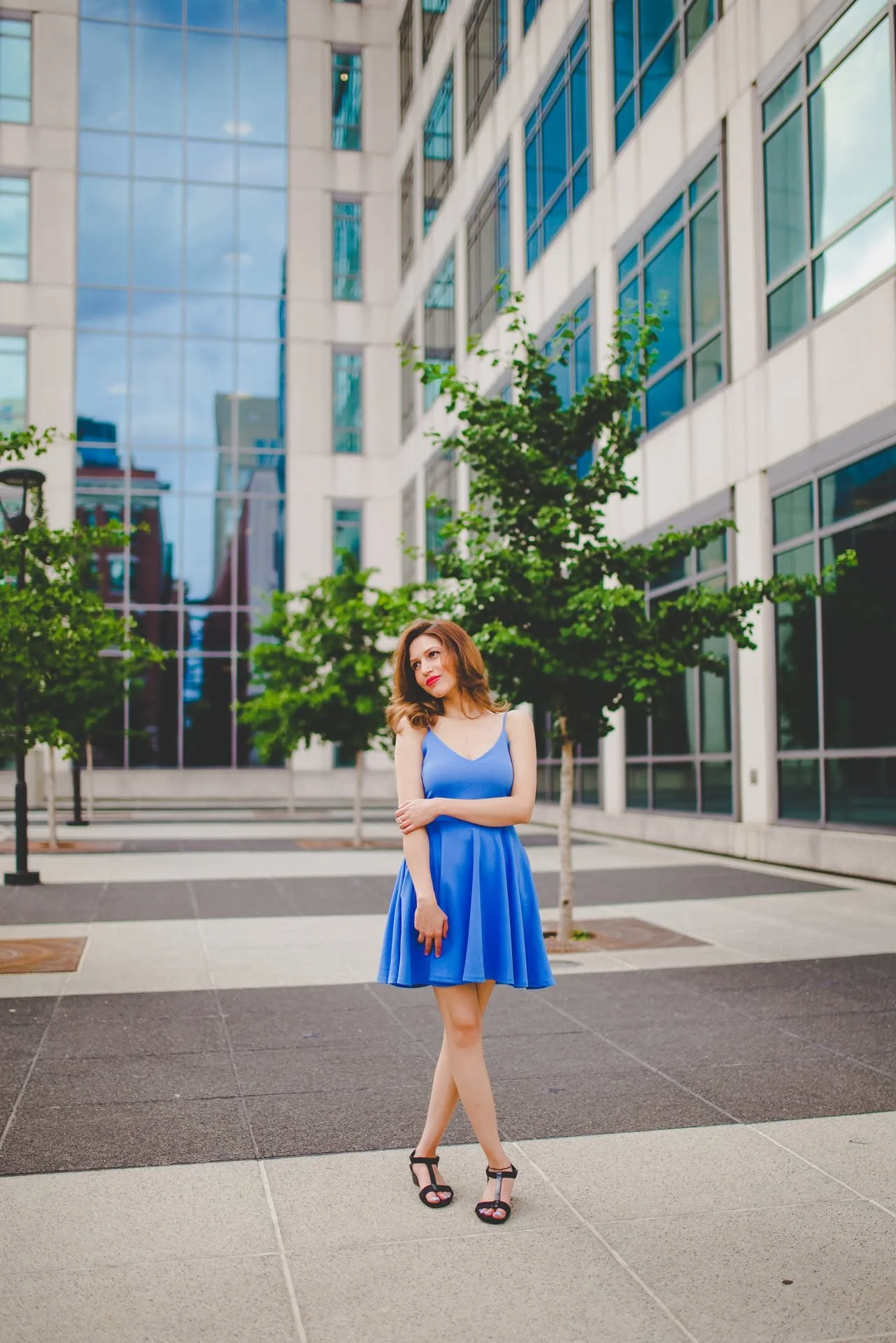 A woman with shoulder-length brown hair and light skin wearing a blue sleeveless dress and black high heels stands outdoors in front of a modern glass building with trees and a paved walkway.