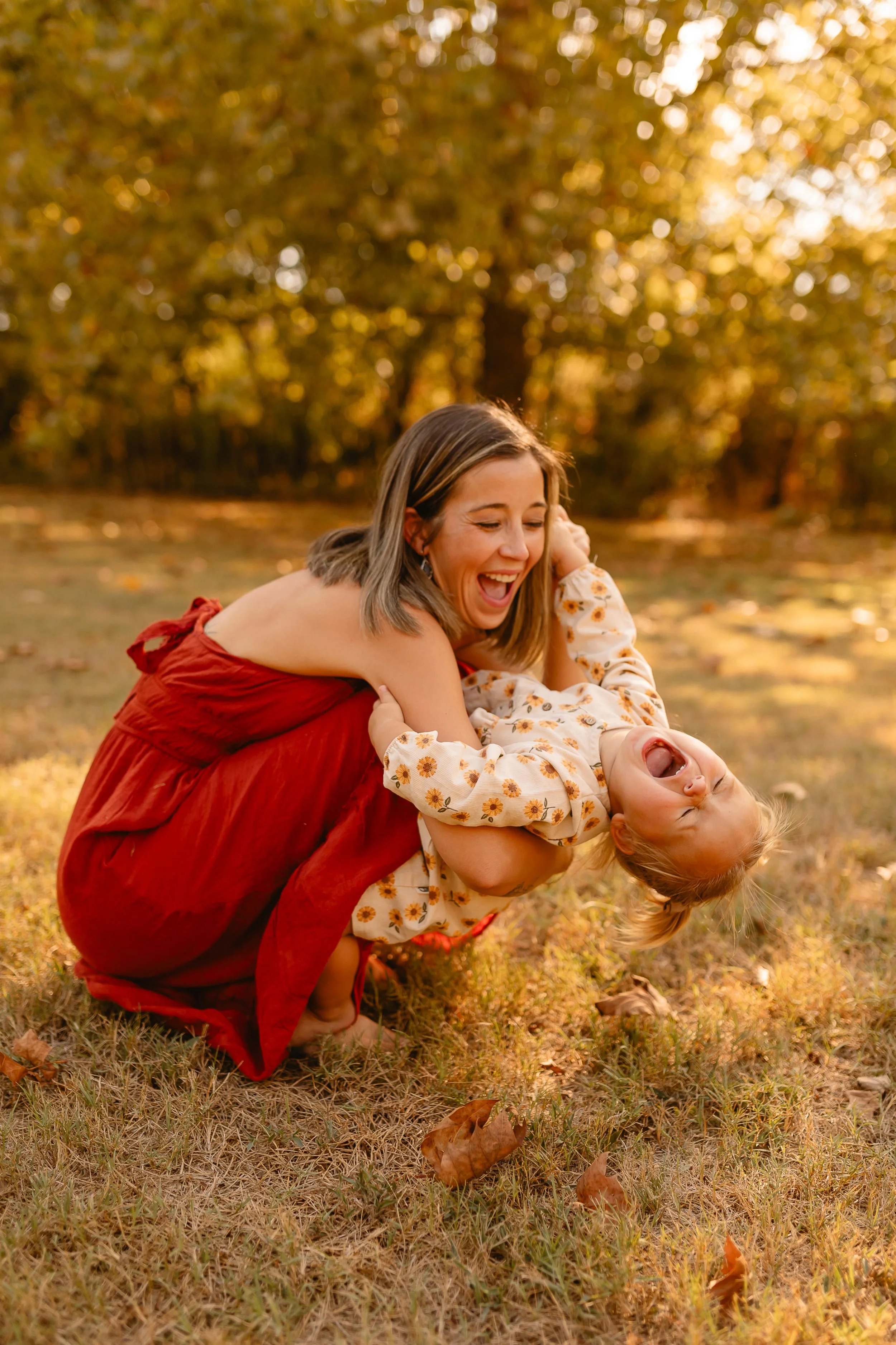 A woman in a red dress is playing with a young girl in an autumn park during sunset. The girl is upside down, laughing and dressed in a floral dress. The background has trees with orange and yellow leaves, and the ground is covered with fallen leaves