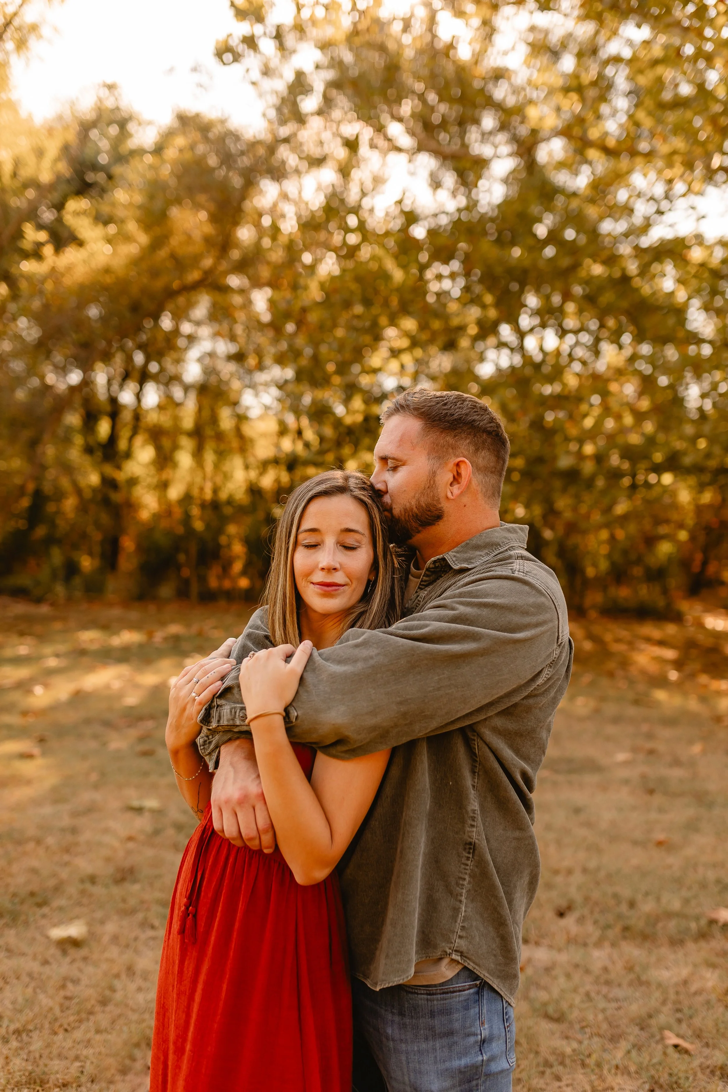 A man and woman hugging outdoors with fall foliage in the background.