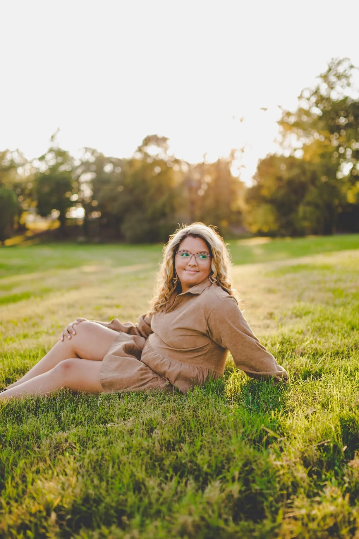 A young woman with curly blonde hair and glasses sitting on green grass in a park during sunset, wearing a brown dress, smiling and looking to the side.