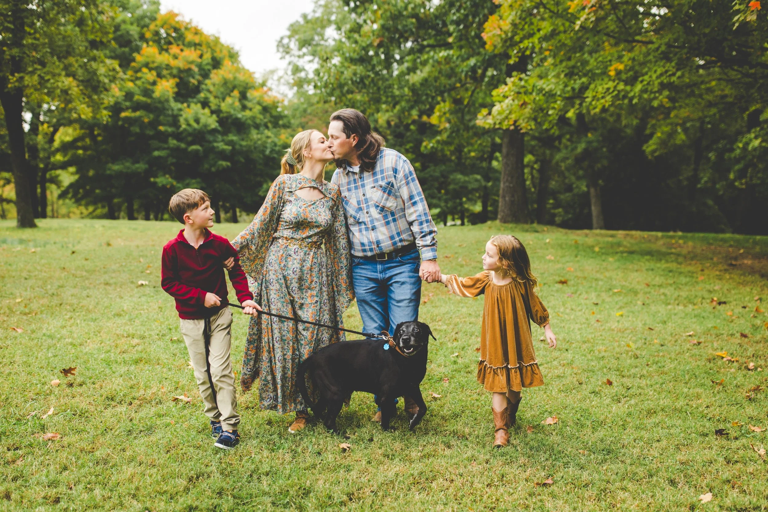 A family of five, including a man, a woman, two children, and a dog, standing on a grassy field with trees showing fall foliage, sharing a kiss.