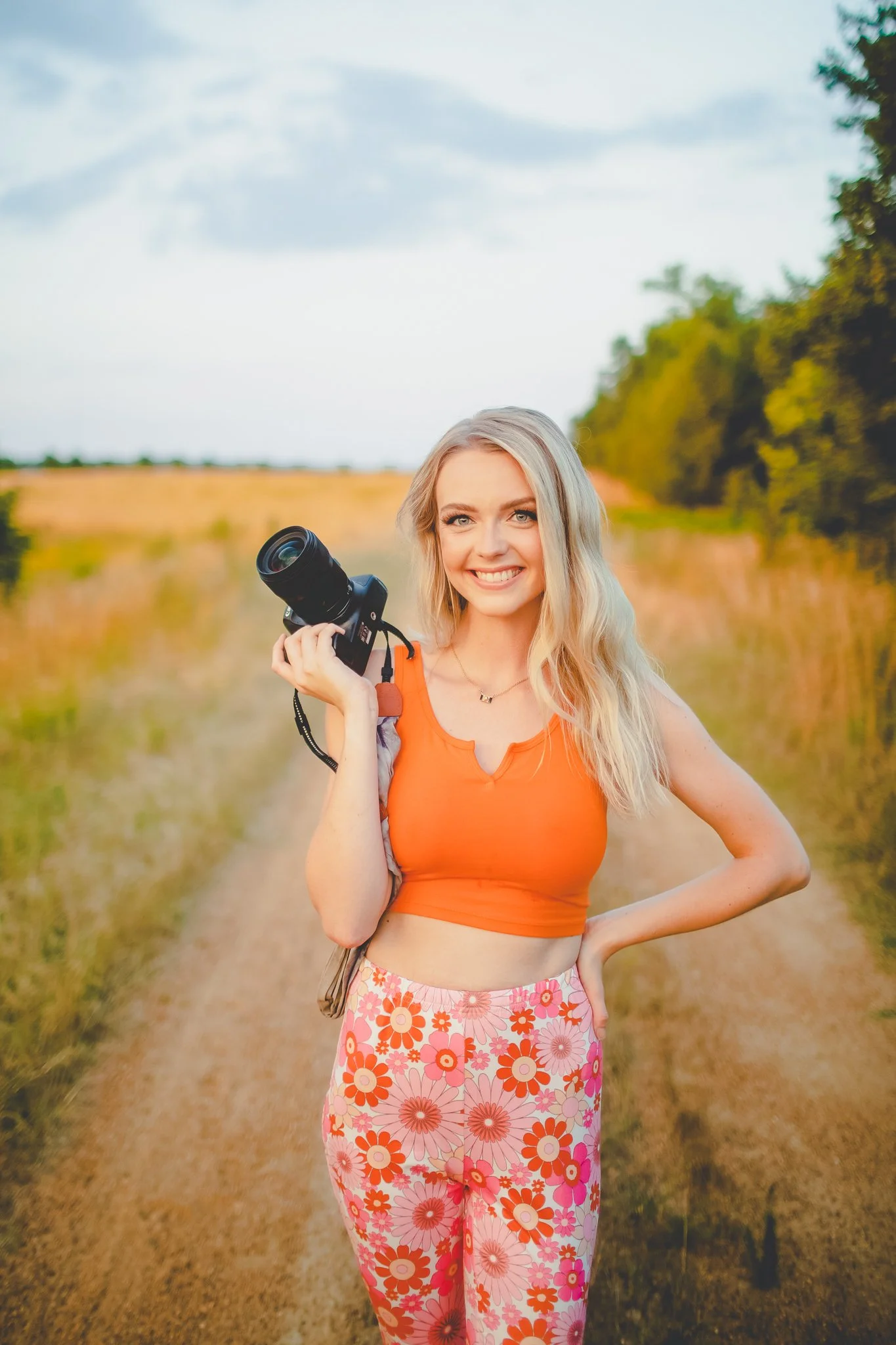 A young woman with long blonde hair smiling and holding a camera on a countryside dirt path with green trees and open field in the background.