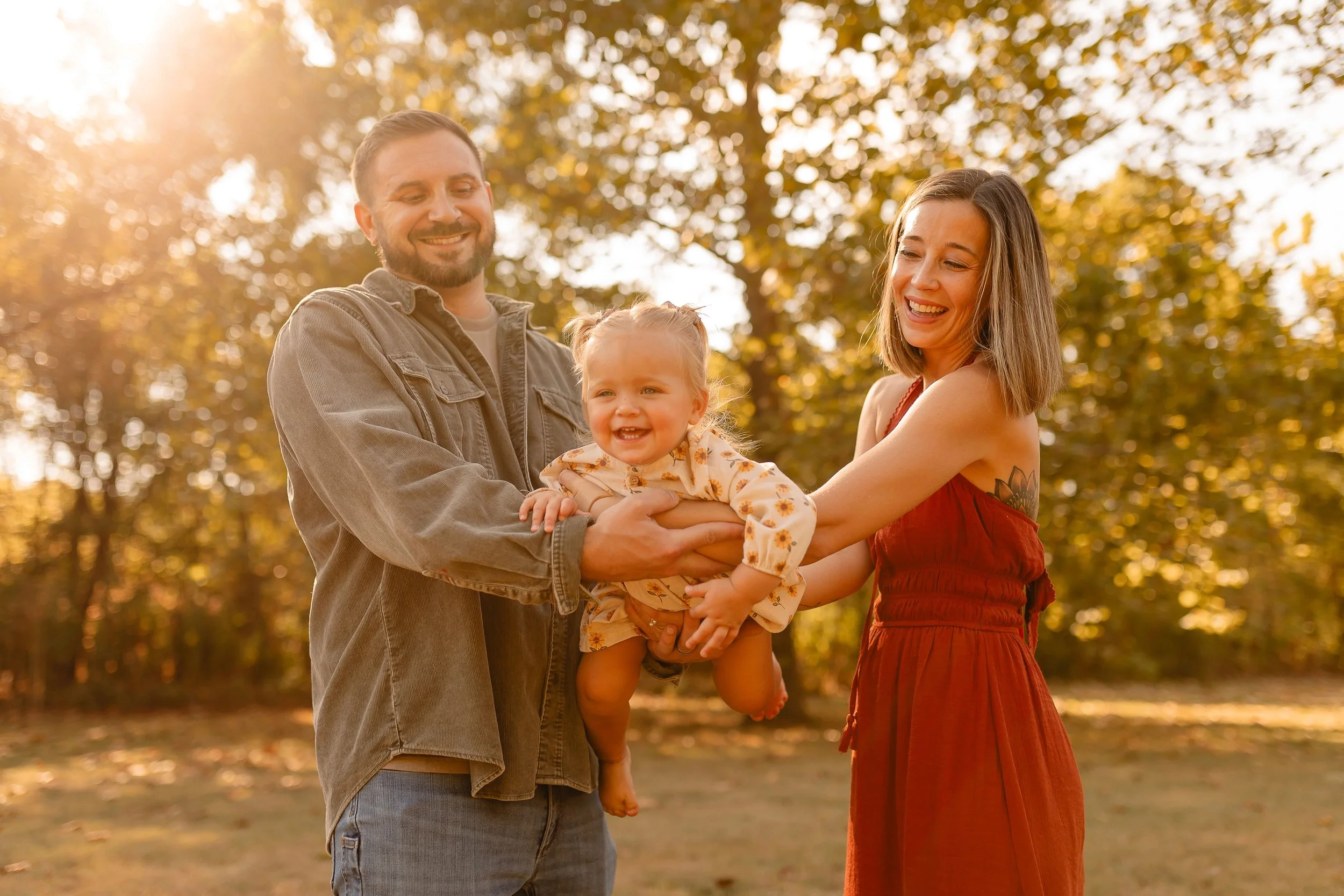 A family of three, a man, a woman, and a young girl, smiling and holding a young girl in a park during autumn with sunlight filtering through trees.