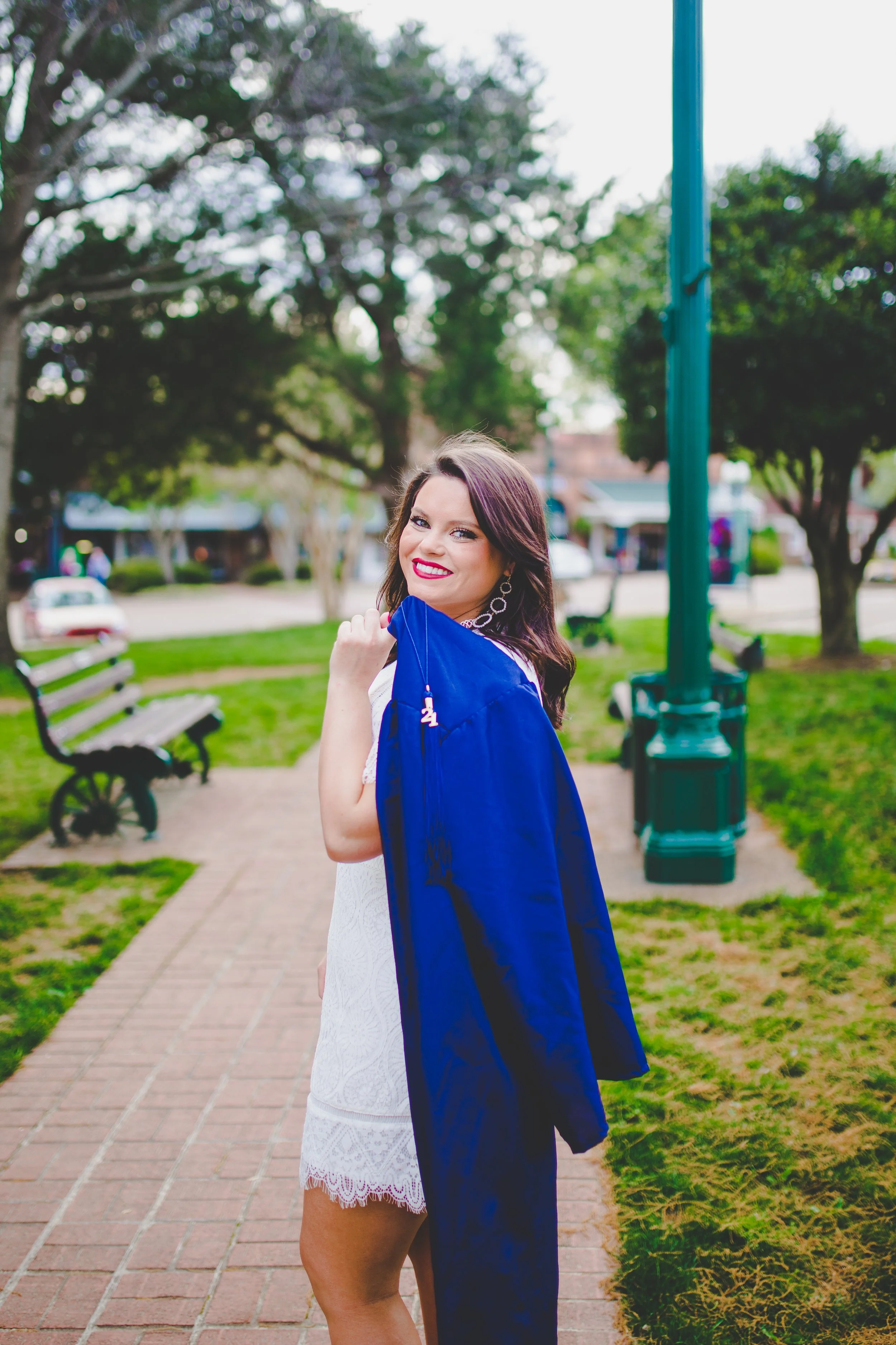 A young woman with dark hair and bright lipstick standing outdoors in a park, wearing a white dress and a blue graduation gown, smiling at the camera. There are park benches and trees in the background.