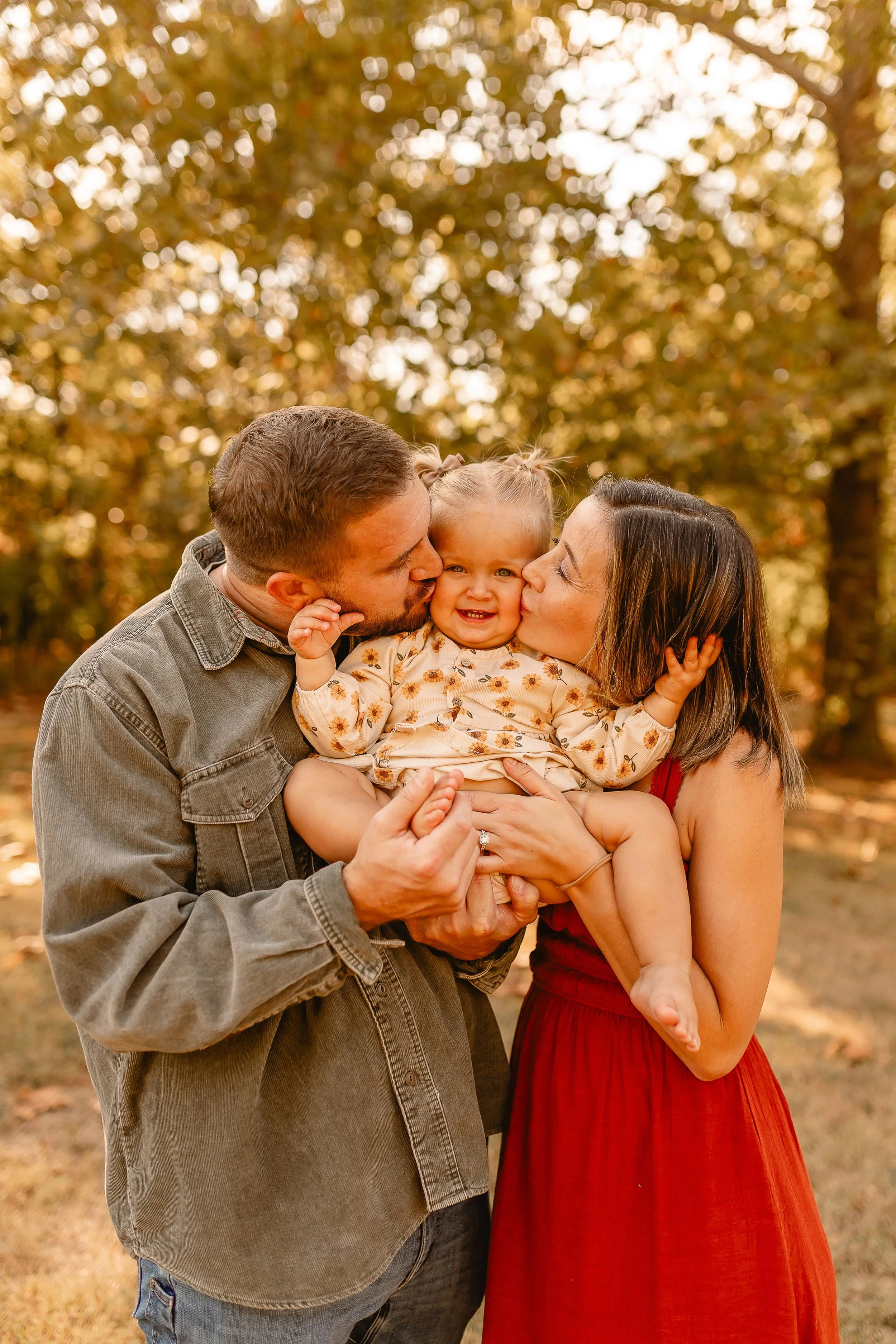 A family of three, consisting of a man, woman, and a young girl, sharing a loving moment outdoors during autumn. The man and woman are kissing the girl on her cheeks while she smiles happily, all surrounded by fall foliage.