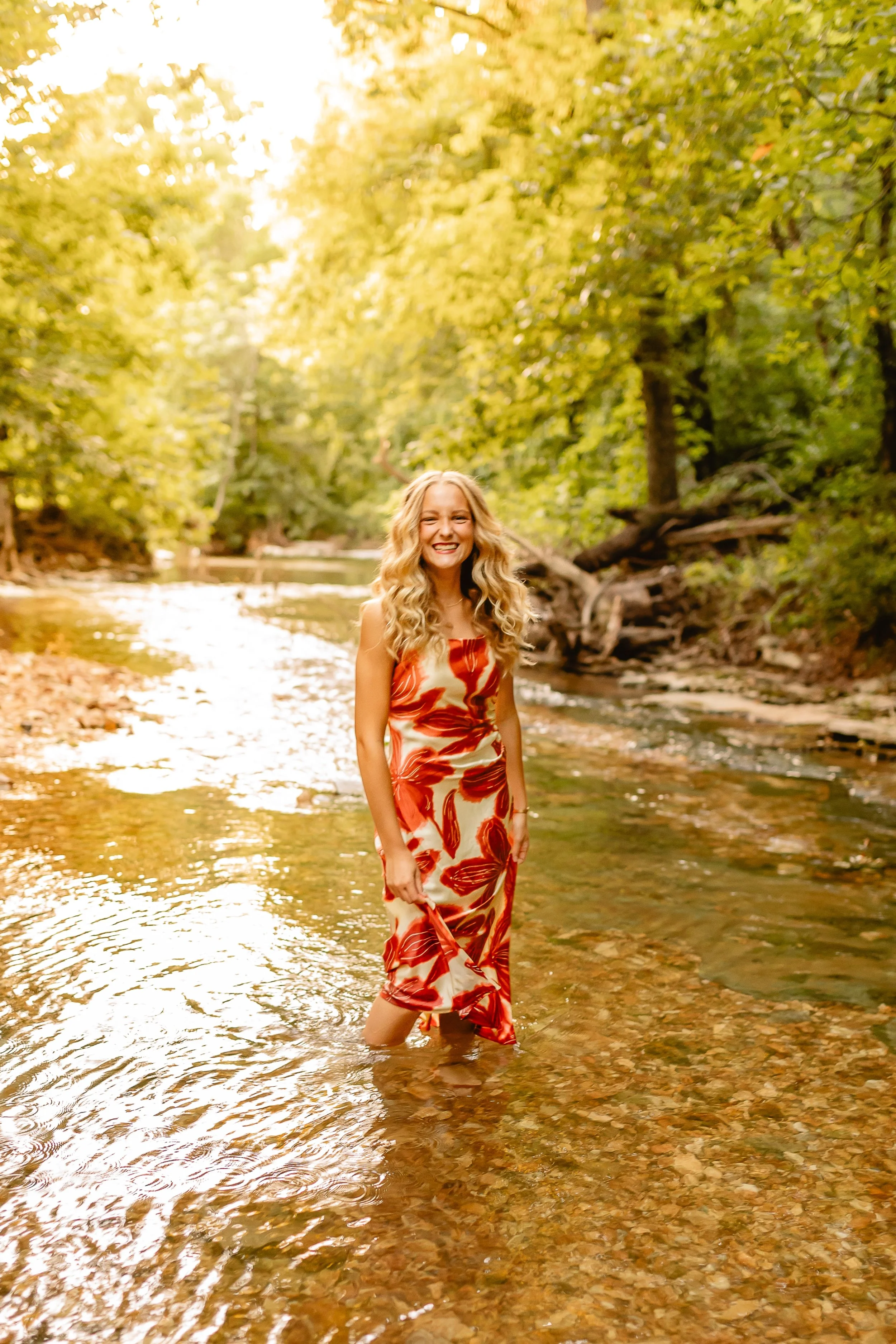 A woman with long, curly blonde hair wearing a red and white floral dress standing in a shallow stream surrounded by trees with green leaves, sunlight filtering through the foliage.