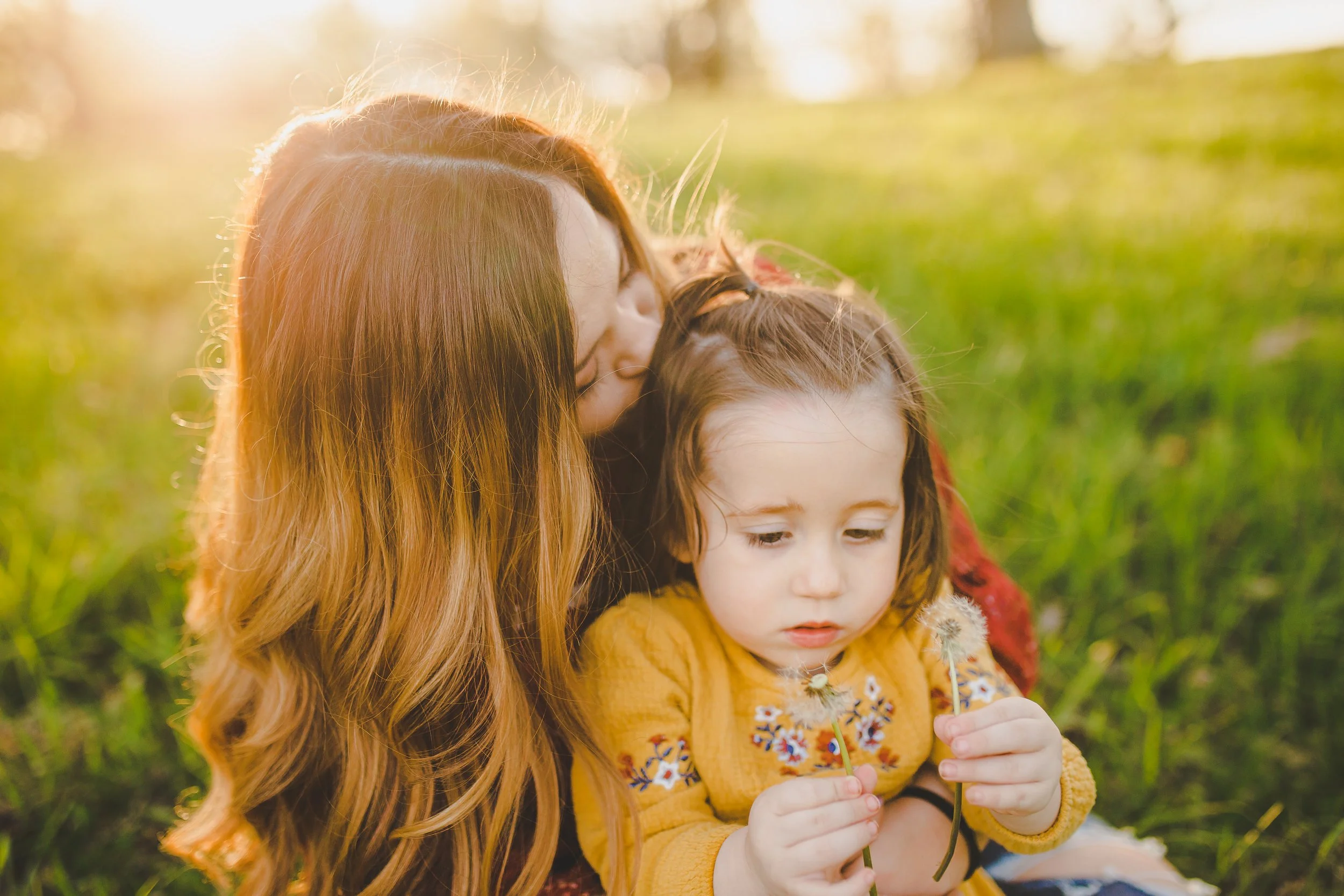 A woman with long red hair and a young girl with brown hair sit outdoors at sunset or sunrise, holding flowers and enjoying a tender moment together.