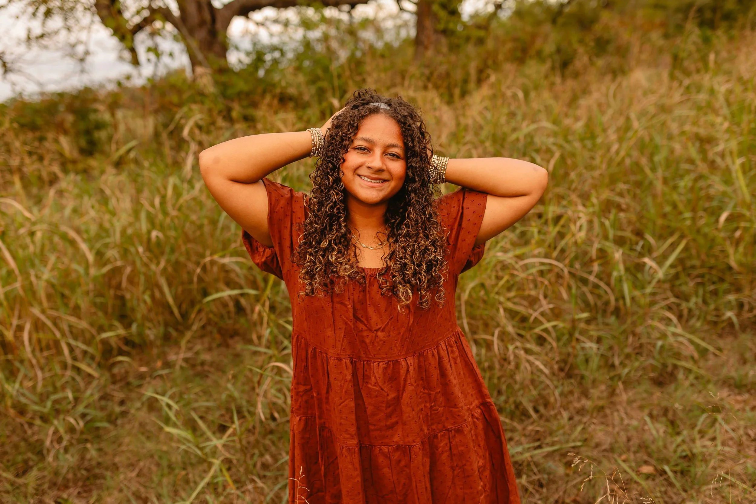 A smiling woman with long curly hair, wearing a rust-colored dress and bracelets, standing outdoors in a grassy field with trees in the background.