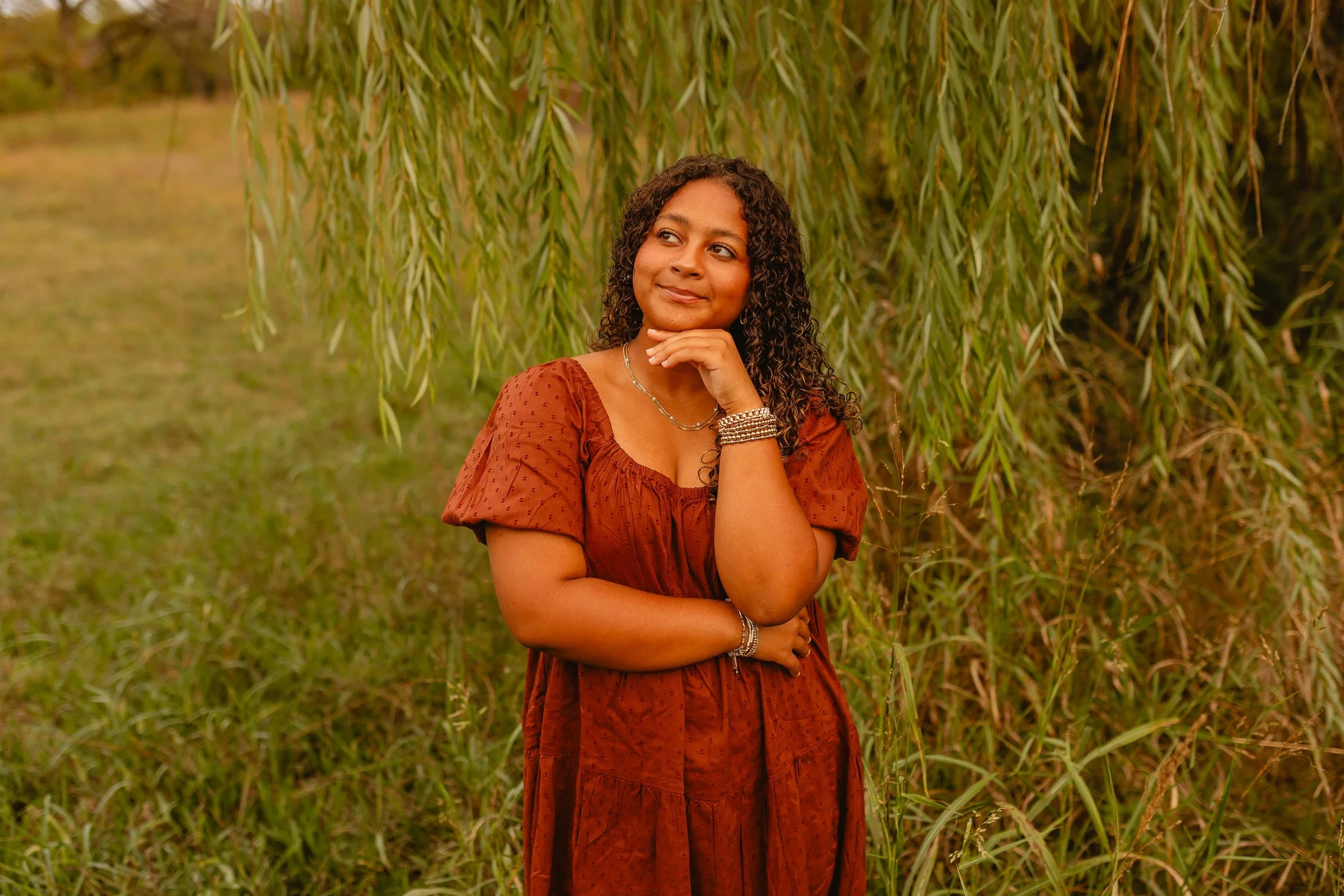 A woman with curly hair wearing a rust-colored dress, jewelry, and a watch, standing outdoors in front of a leafy tree with green grass below, smiling softly.