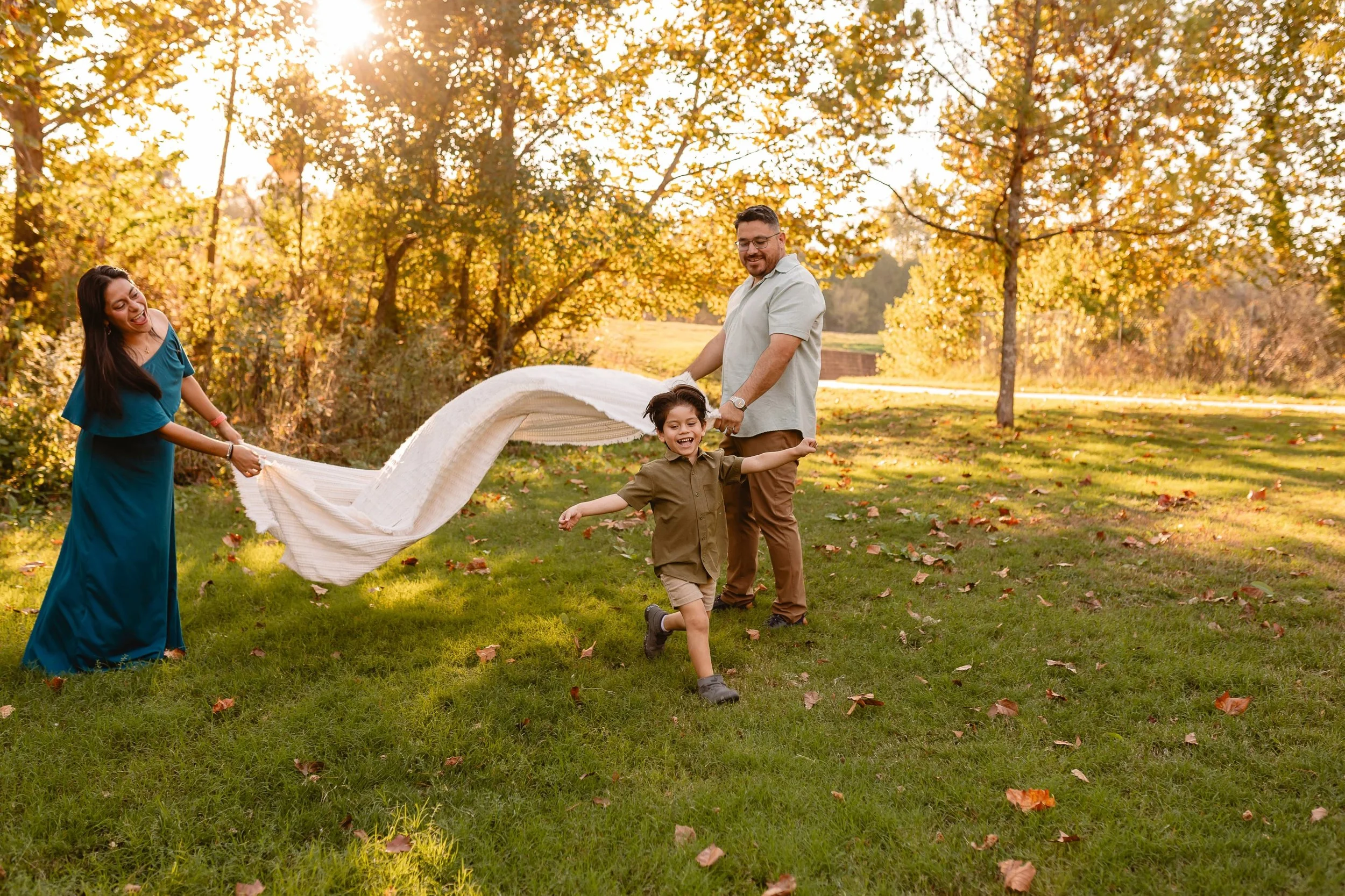 A family of four playing outdoors in a park during fall, with vibrant autumn leaves and sunshine in the background. The mother and father hold a blanket while their children run around happily.