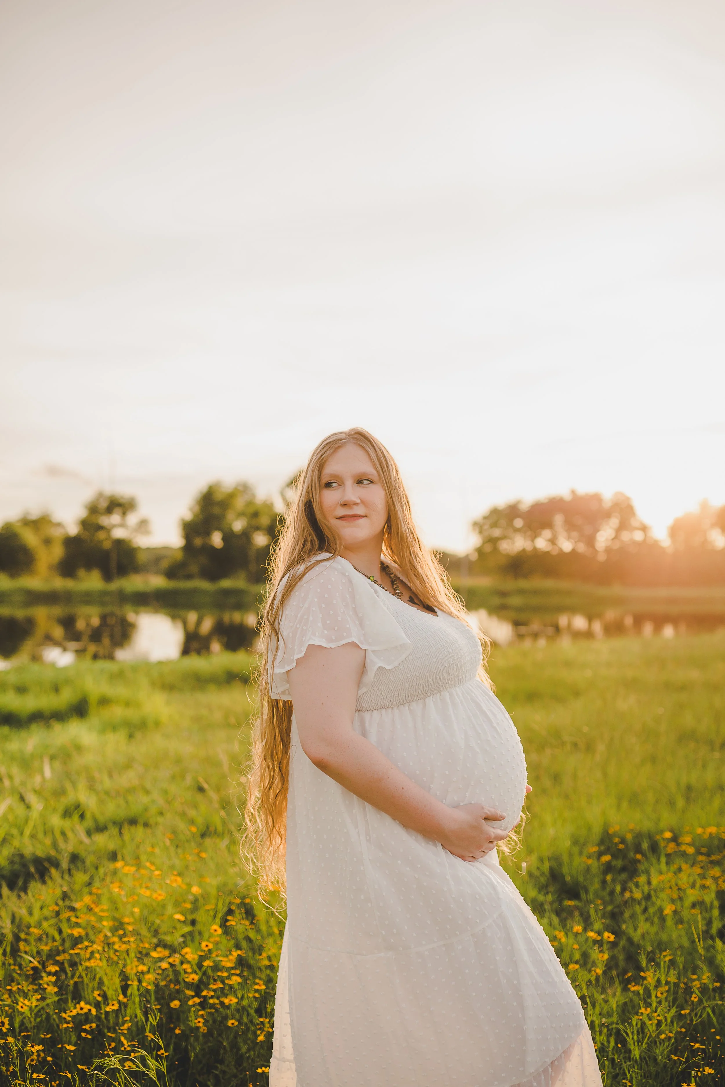 Pregnant woman with long hair in white dress standing in a field of yellow flowers at sunset.