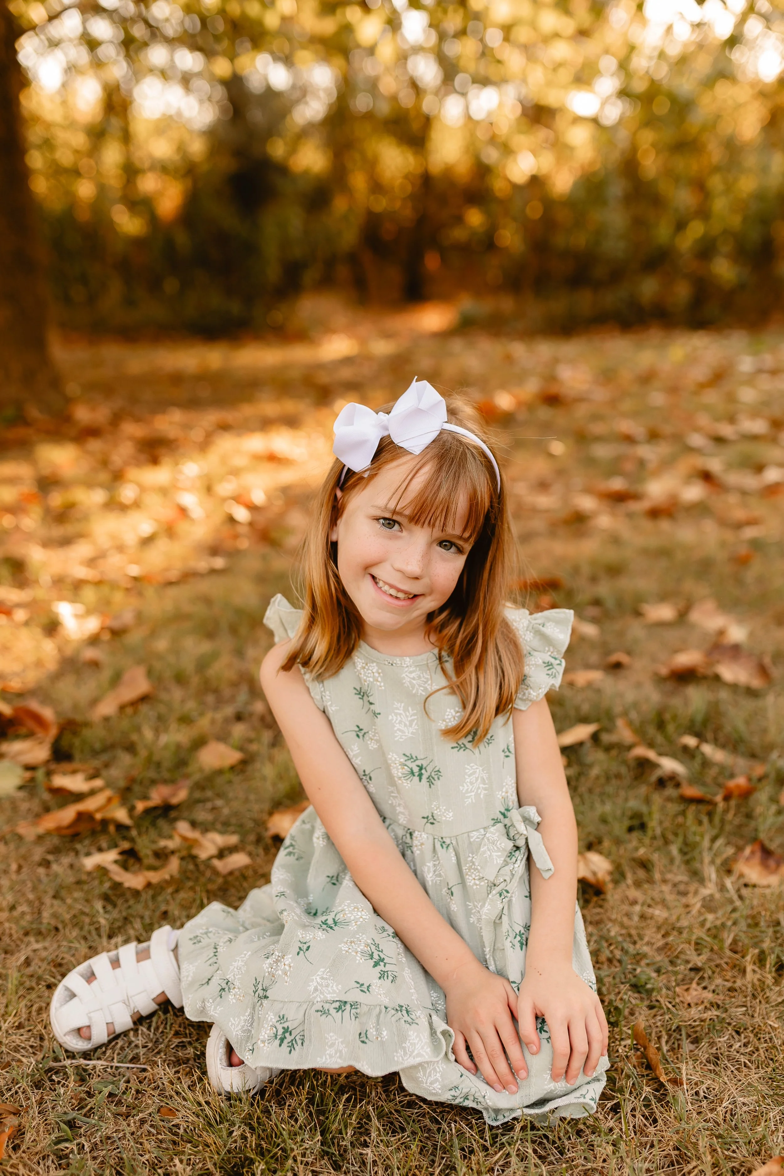 A young girl with red hair, wearing a white bow headband and a light-colored dress with green floral embroidery, sitting on the grass amidst fallen autumn leaves, smiling at the camera.