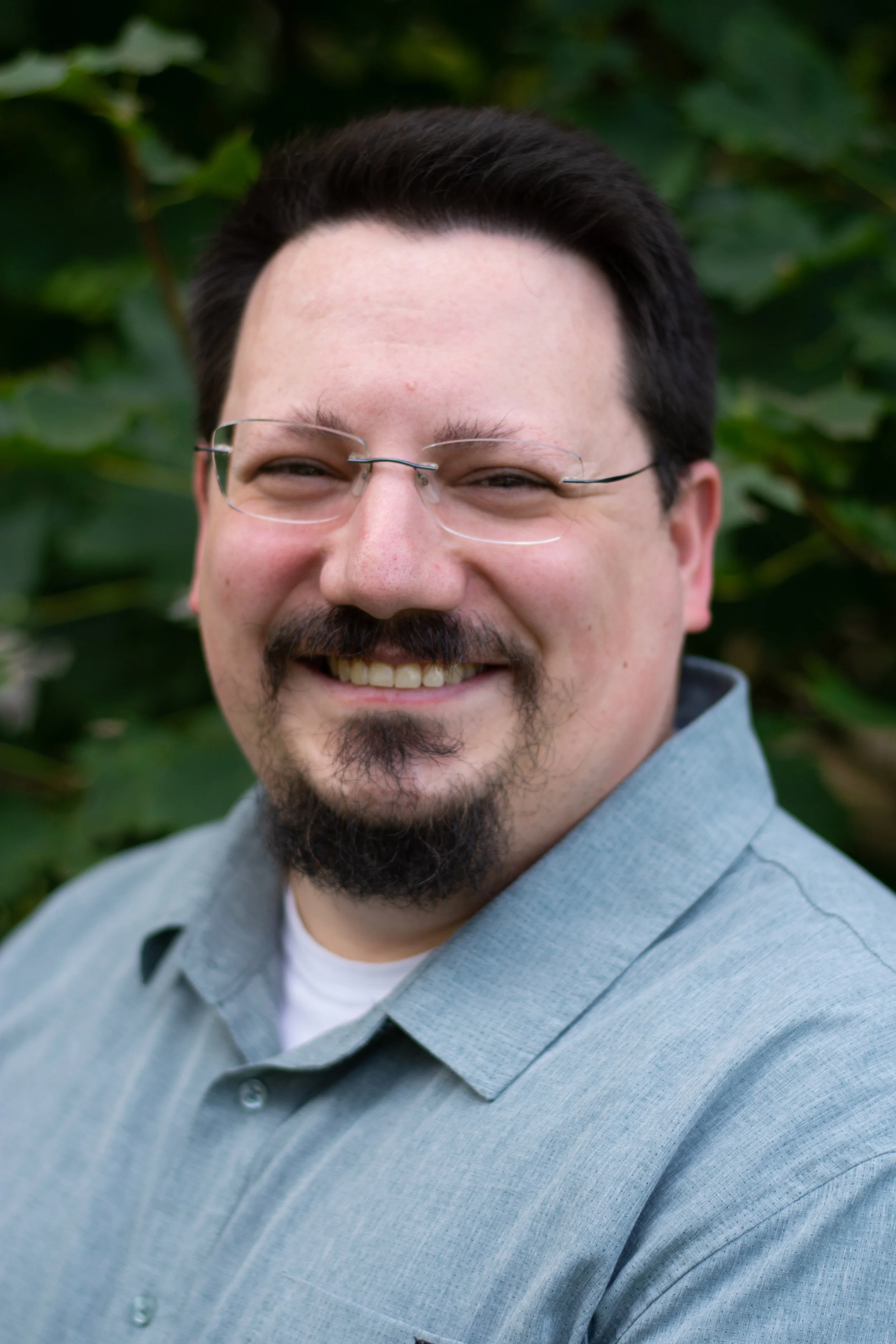 A smiling man with glasses, dark hair, and a goatee, wearing a gray collared shirt, standing outdoors with blurred green foliage in the background.