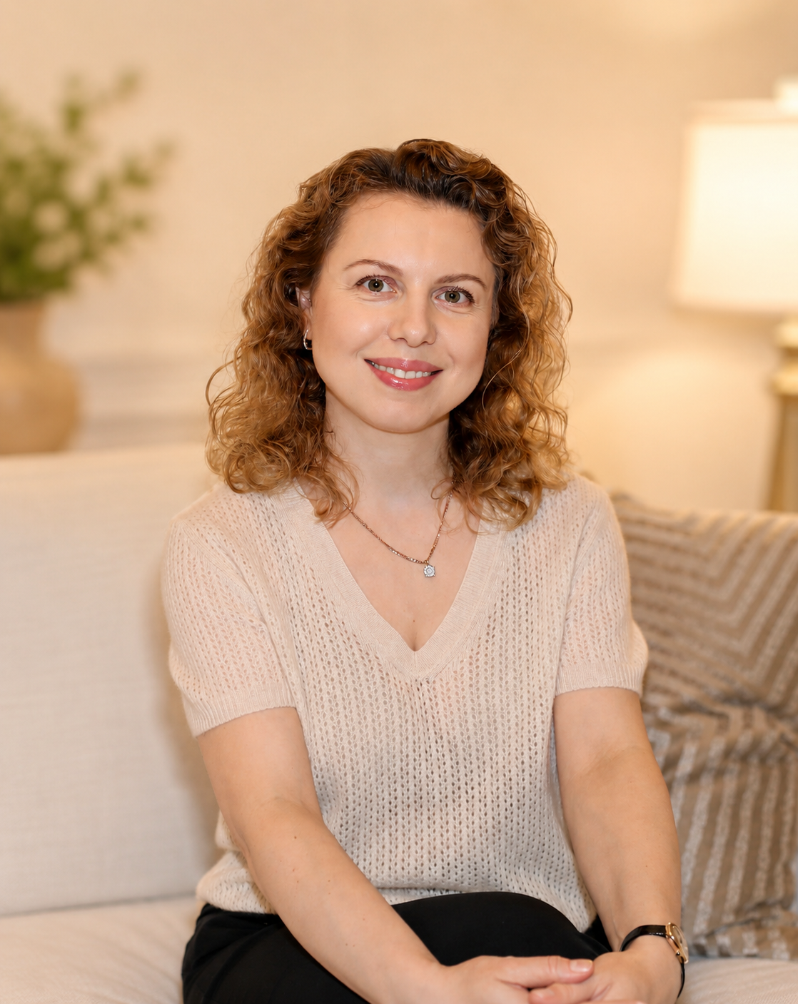 A woman with curly red hair, wearing a beige sweater and silver jewelry, sitting on a beige couch in a cozy living room with warm lighting and a lamp in the background.