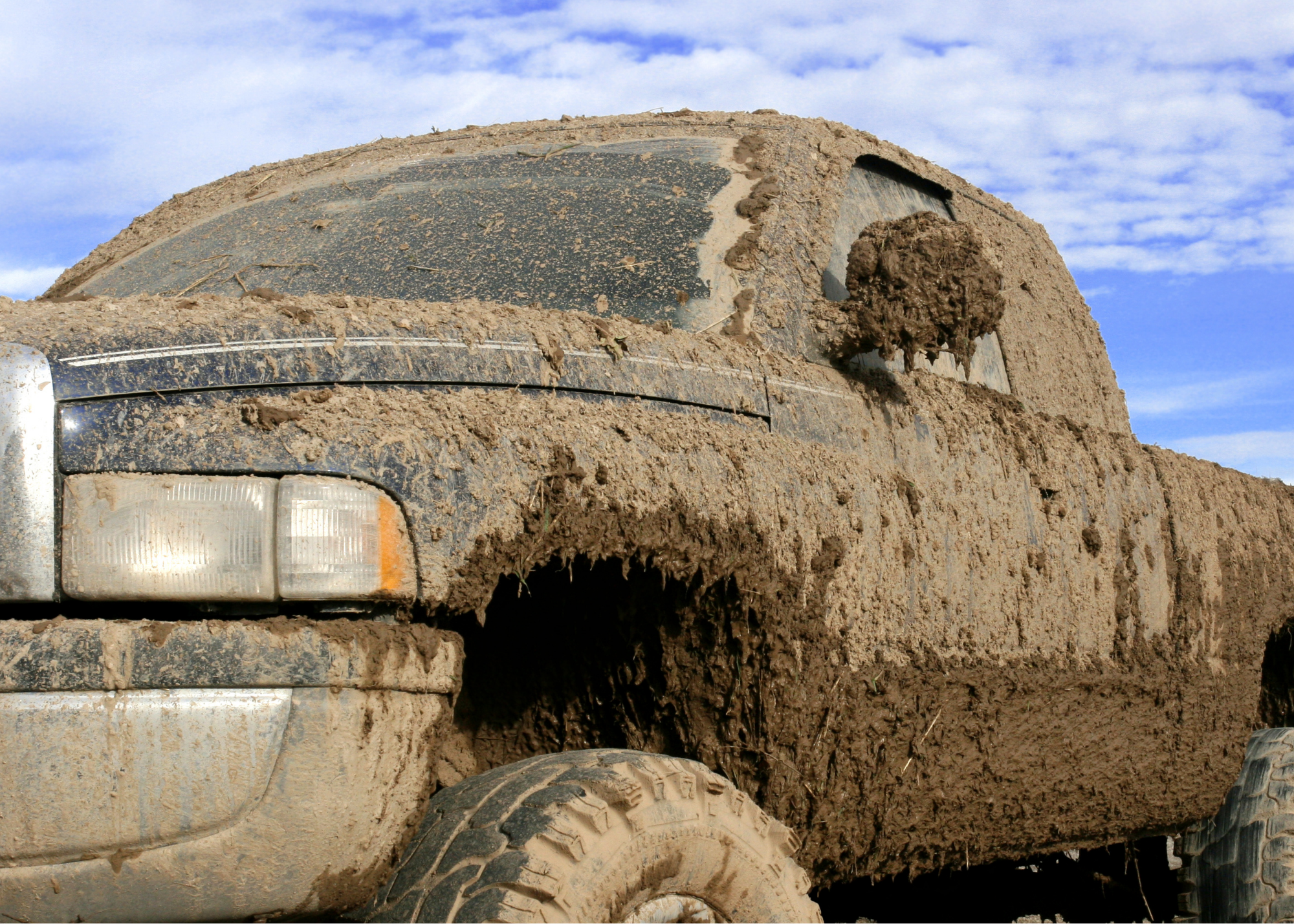 Close-up of the front of an off-road vehicle covered in mud, with visible headlights and tire, against a blue partly cloudy sky.
