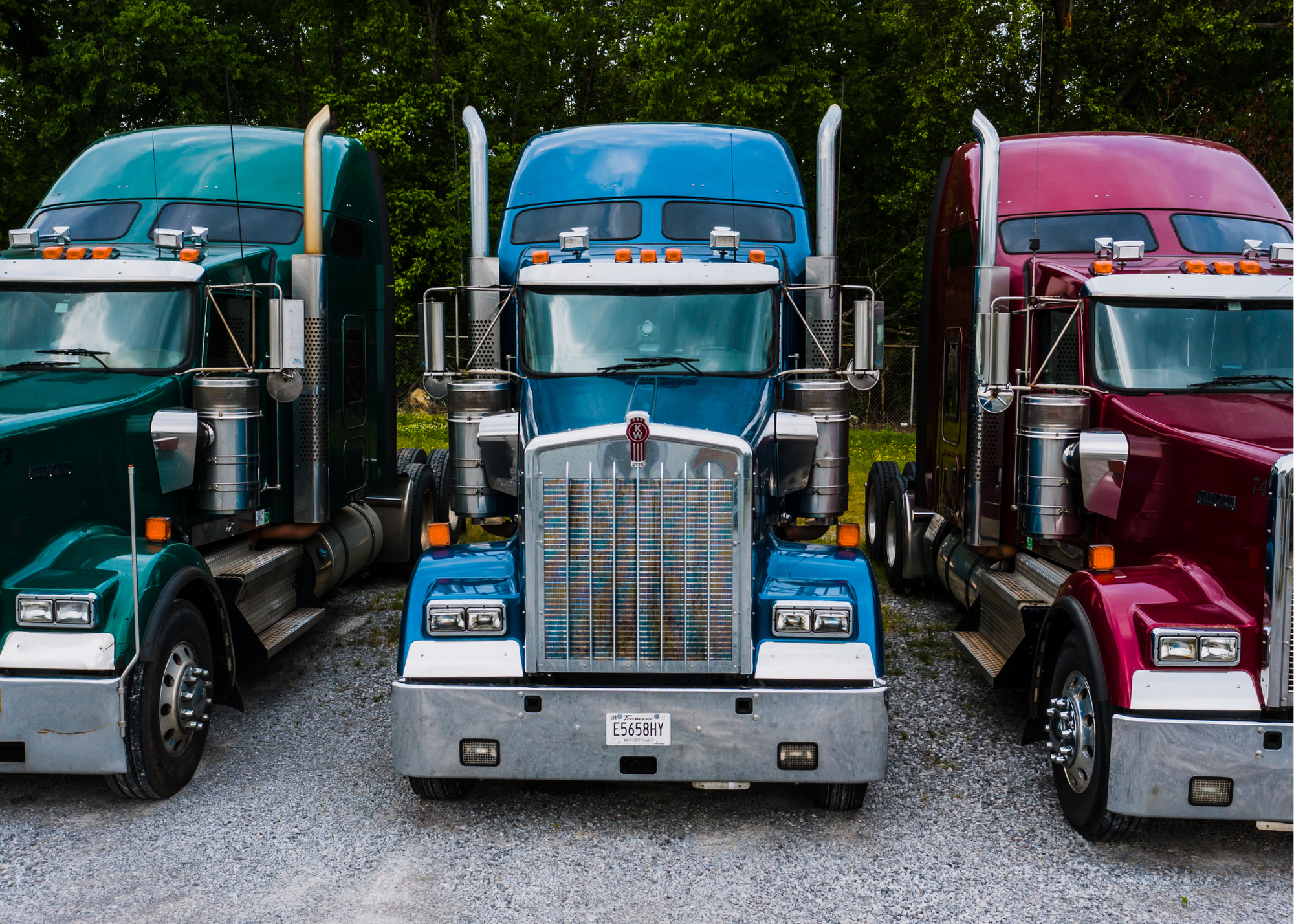 Front view of three large semi-trucks parked side by side on a gravel surface with trees in the background. The trucks are green, blue, and red. The trucks have large grilles, headlights, and chrome details.