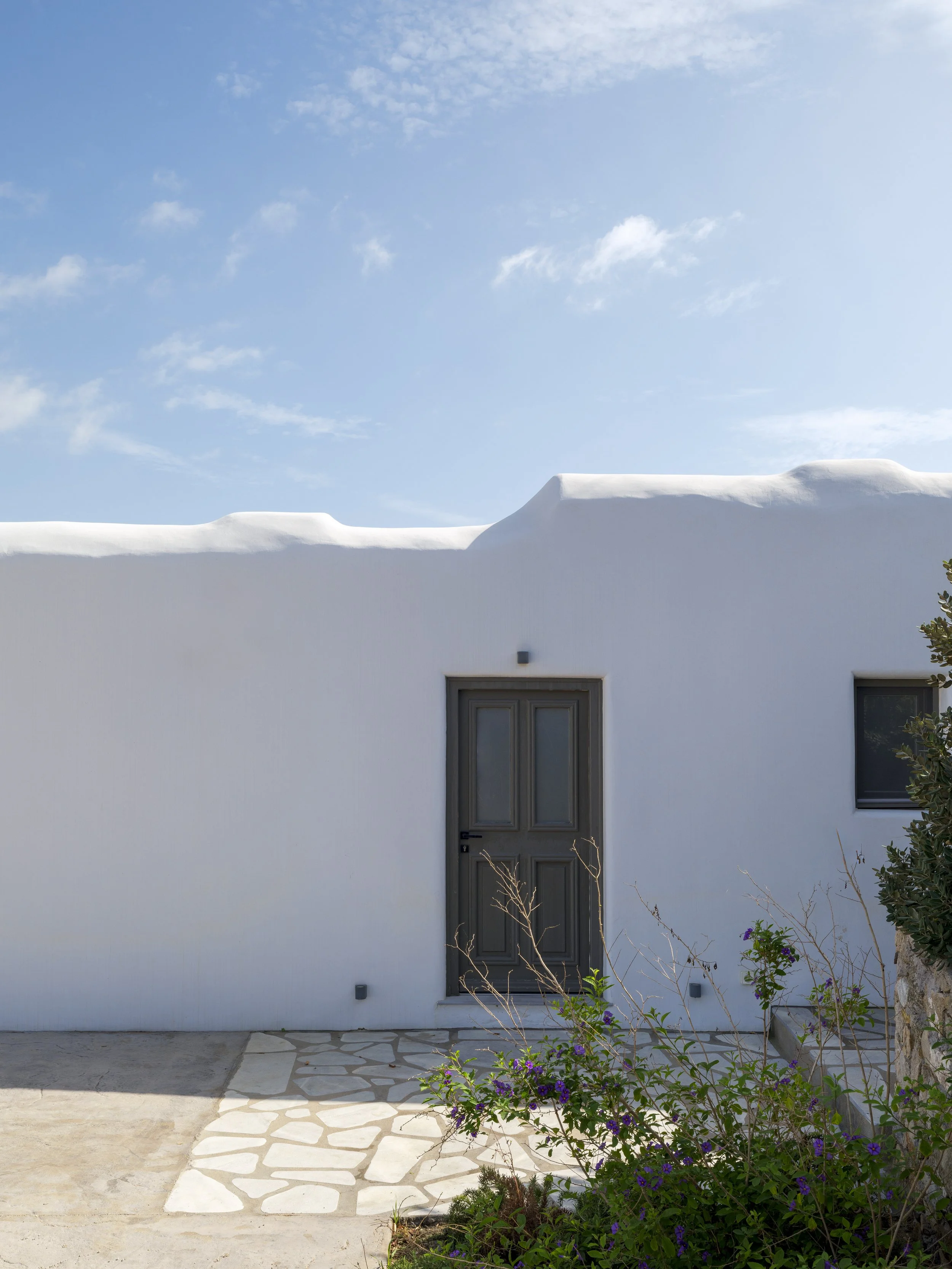 White building with a flat roof, gray door, and small windows. Some plants and flowers are in front of the building, with a paved pathway and blue sky with clouds above.