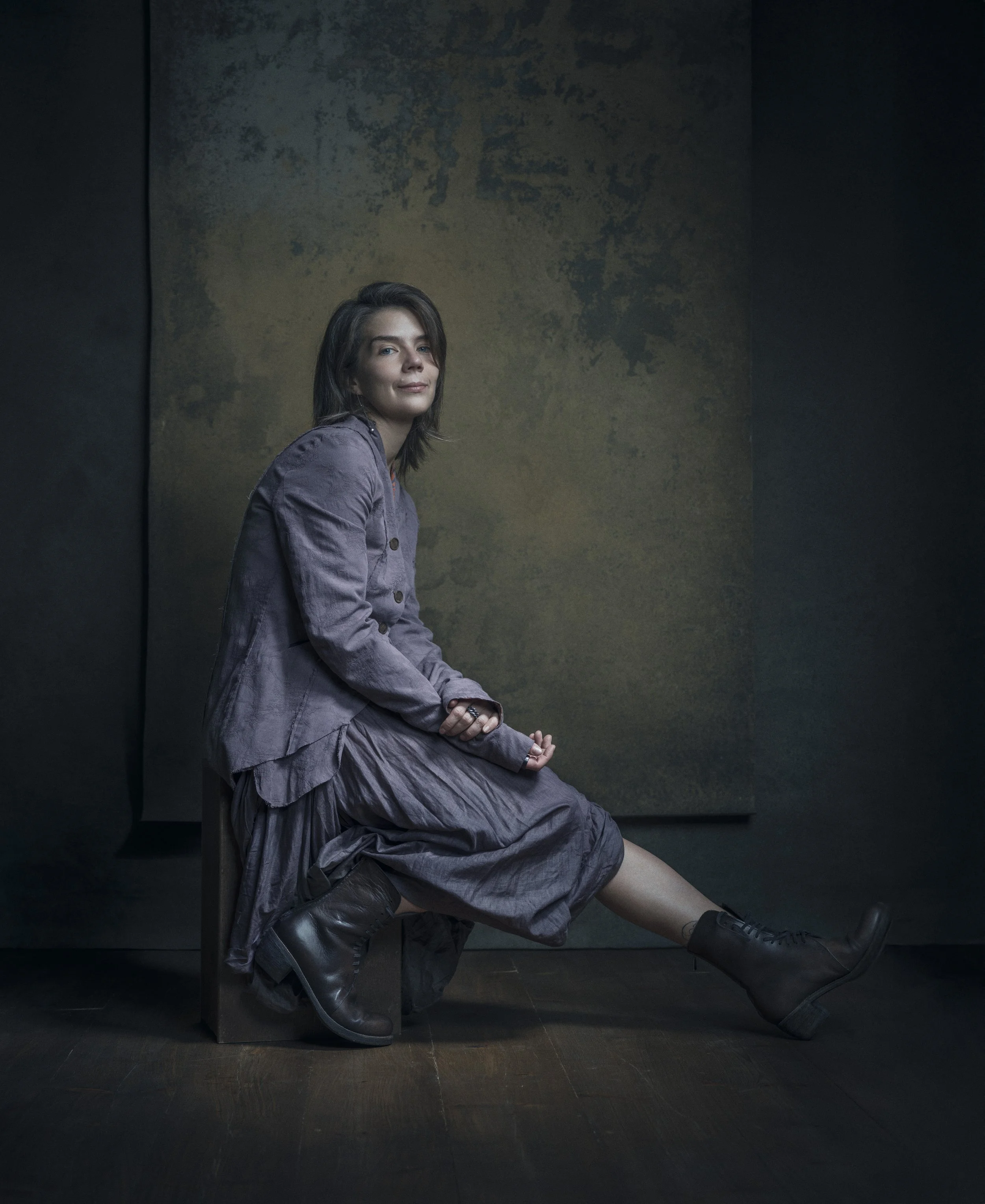 A woman sitting on the floor with her back against a wall, wearing a purple jacket and long skirt, with black boots, in front of a dark wall with faint abstract texture.
