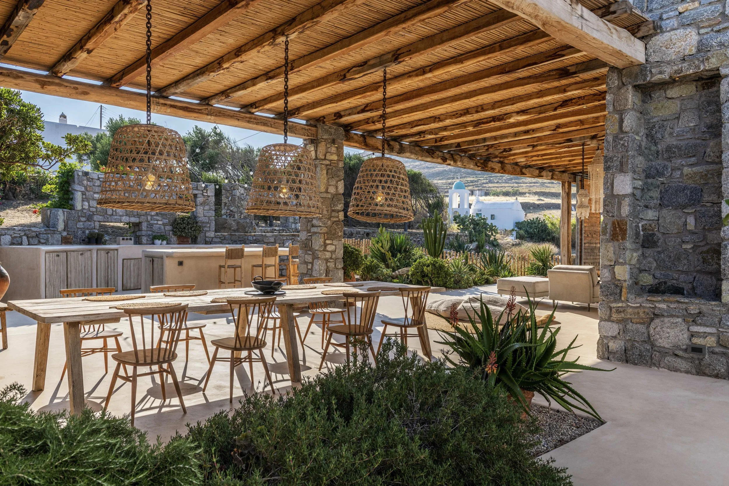 Outdoor patio area with a wooden ceiling, hanging woven rattan light fixtures, stone pillars, a long wooden dining table with chairs, and surrounding desert plants including cacti, with a view of a white building in the distance.