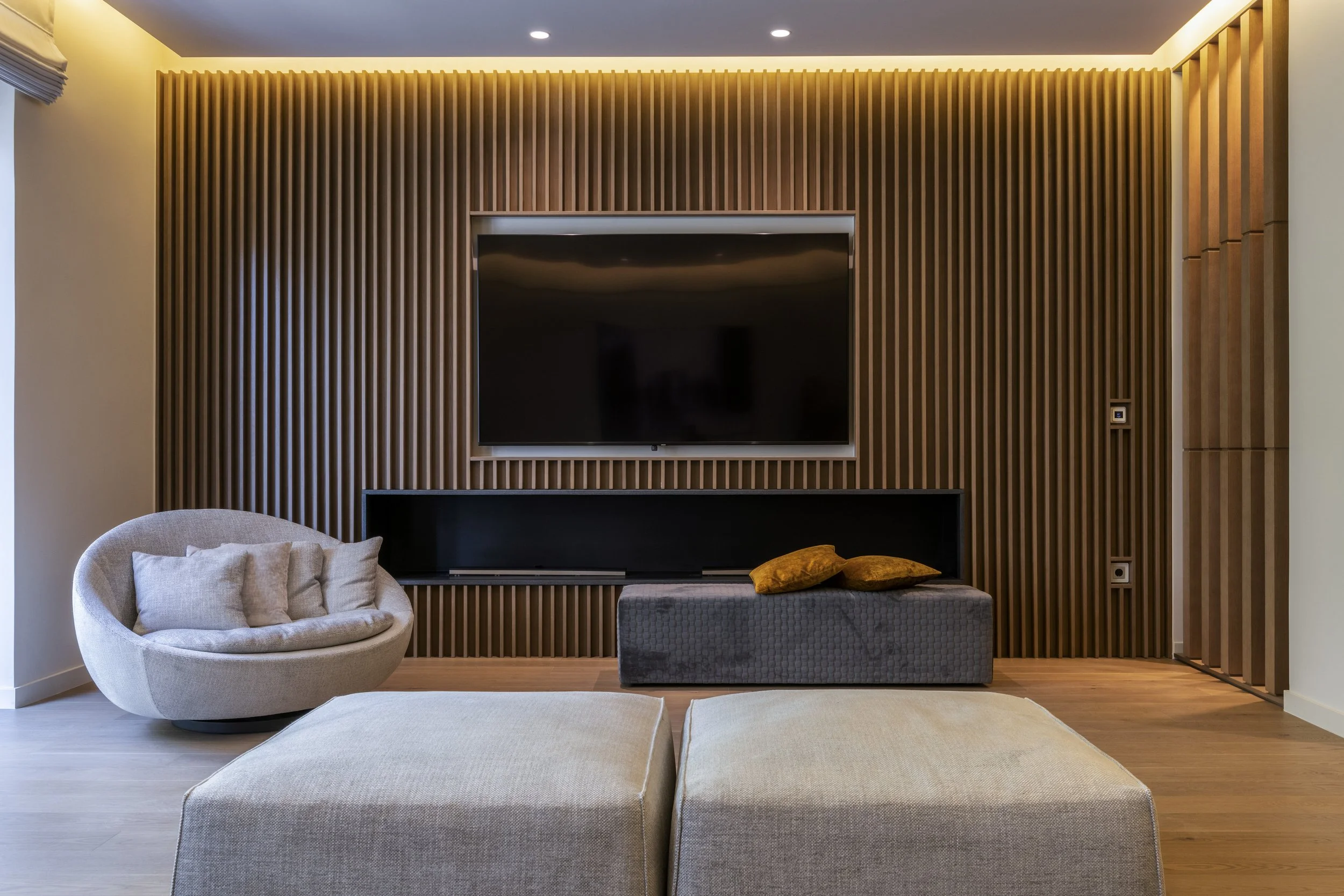 Modern living room with a gray upholstered ottoman, a textured gray bench with two yellow pillows, a beige curved armchair with throw pillows, and a wooden accent wall with a mounted TV. Recessed lighting and a window with a gray blind are visible.
