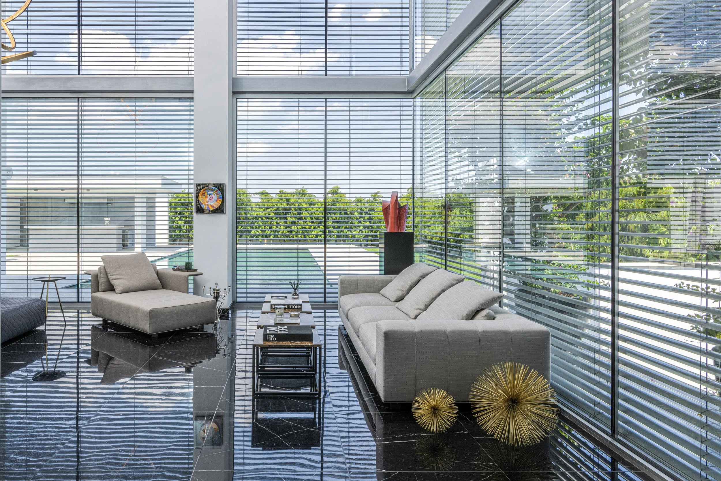 Modern living room with large glass windows and blinds, light-colored sofas, black marble flooring, and outdoor view of greenery.