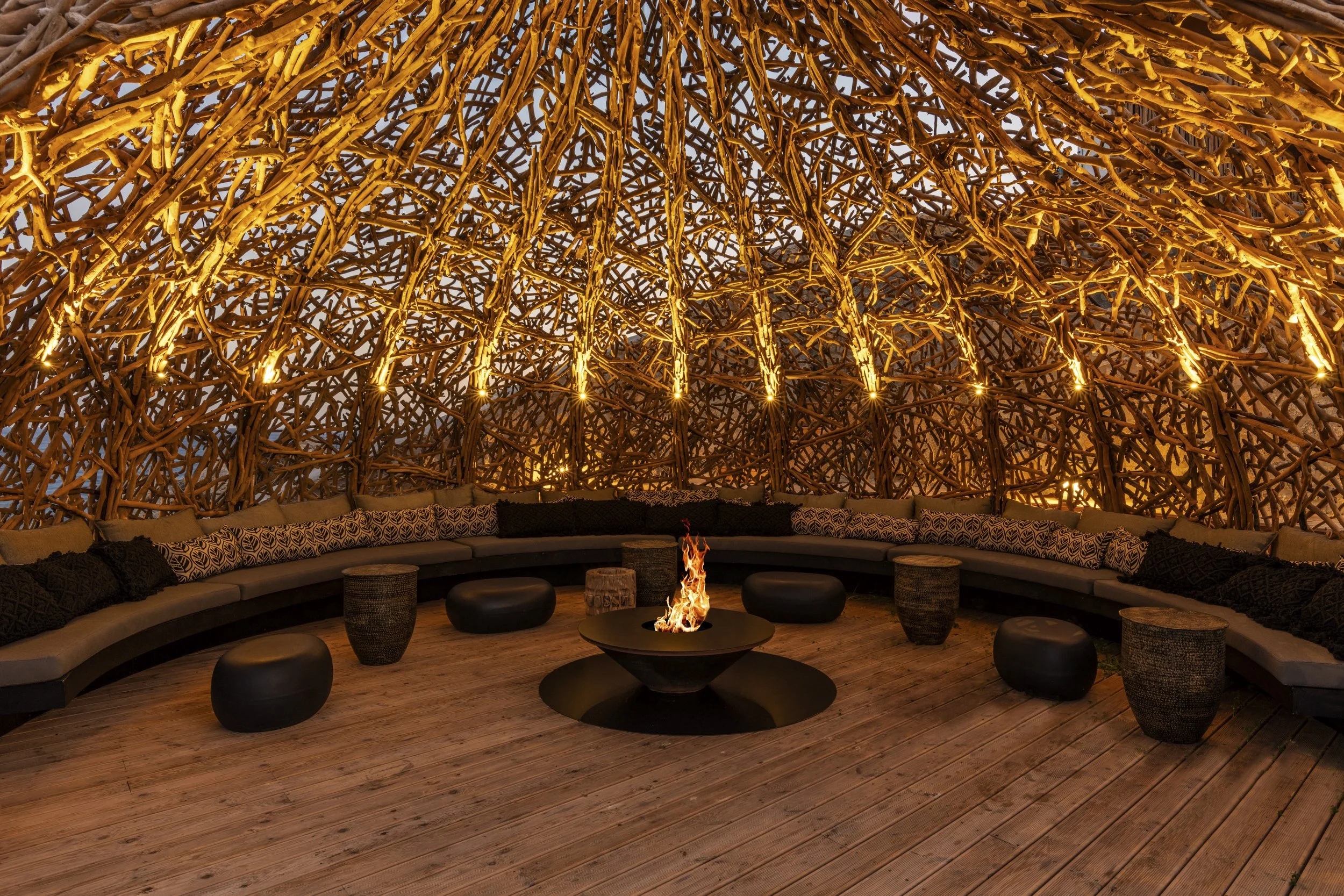 Interior of a circular lounge area with a woven wooden dome ceiling, a fire pit in the center, along with cushioned seating and small tables around it.