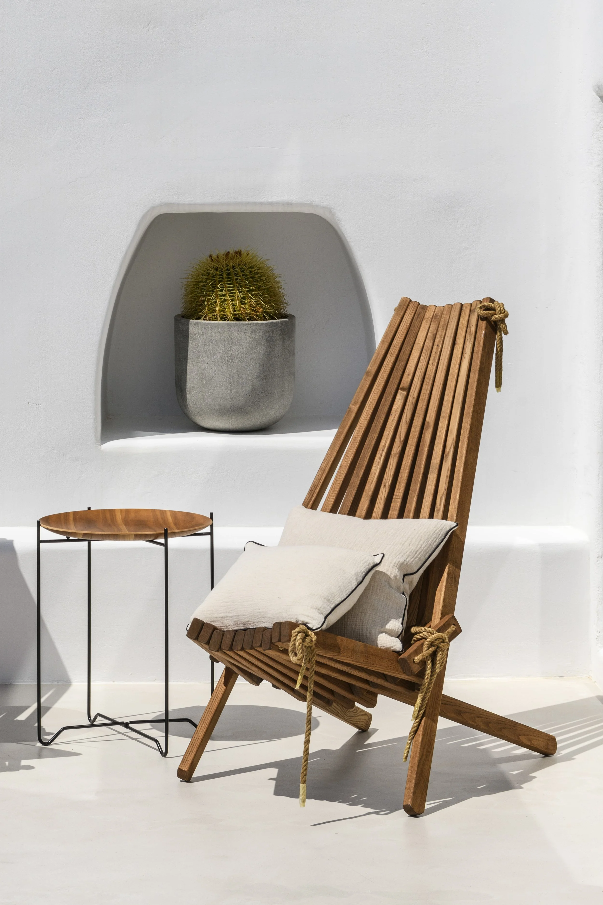 A wooden lounge chair with a cream cushion and pillow, set against a white wall with decorative niches. One niche contains a potted cactus in a gray concrete pot, and a small round side table with a wooden top and black metal legs is beside the chair