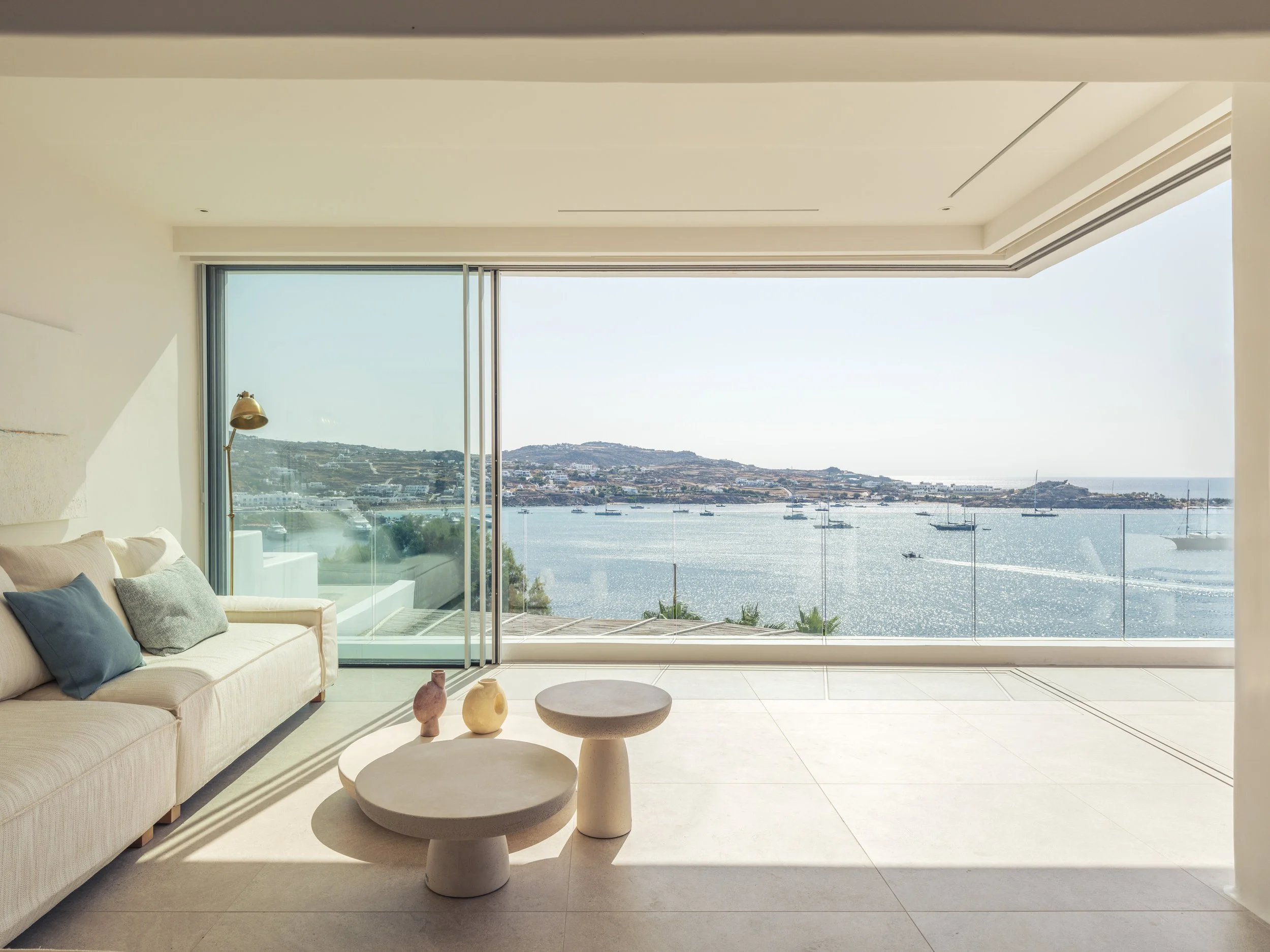 Modern living room with large glass sliding doors opening to a view of a harbor with sailboats, hills, and buildings in the background. Interior features a beige sofa with throw pillows, a floor lamp, and a set of modern, rounded coffee tables with v