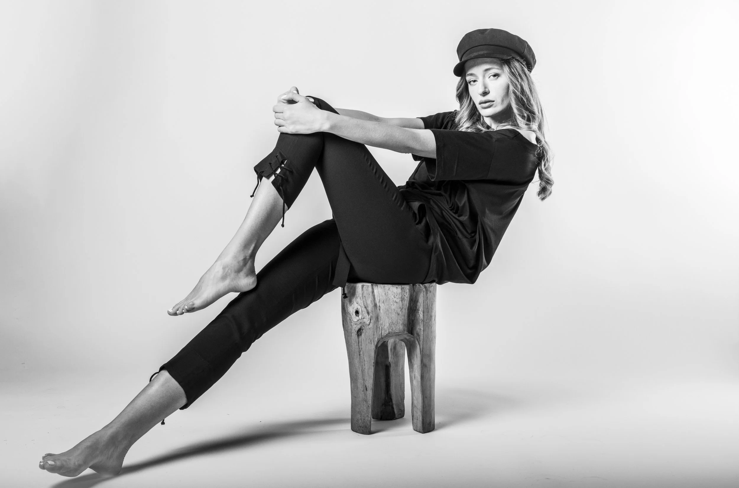 Black and white photo of a woman sitting sideways on a wooden stool, wearing a hat and casual outfit, with her legs extended and her arms resting on her knee, against a plain background.