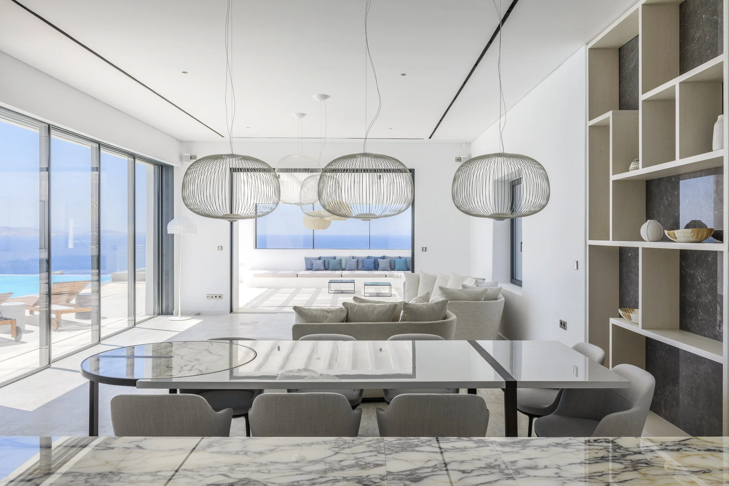 Modern living room with large windows overlooking the ocean, white walls, a white sofa with cushions, hanging pendant lights, and a marble dining table.