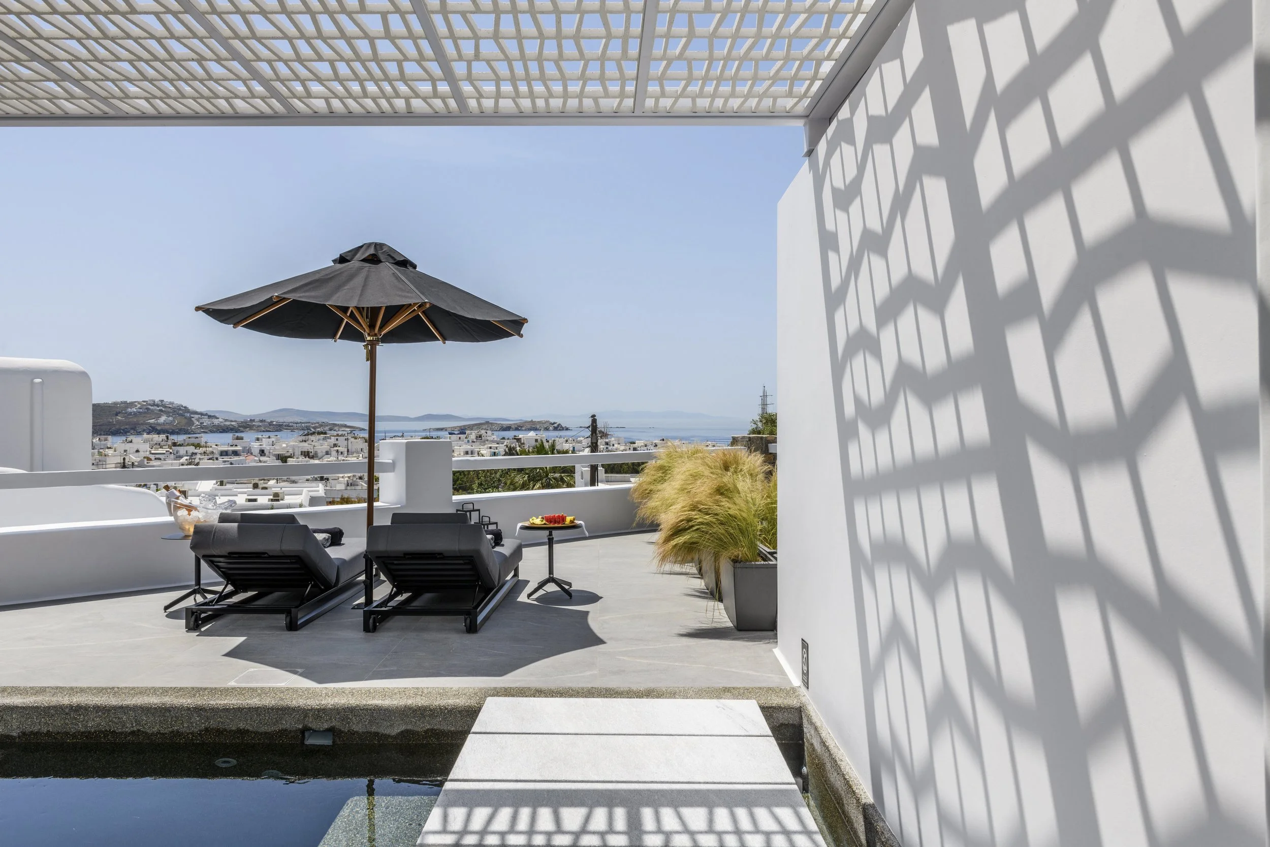 A rooftop patio with black loungers, a black umbrella, and a small table with red and yellow items. The view overlooks white buildings, a hilly landscape, and the ocean with distant islands under a clear sky. White walls and a geometric pergola cast 