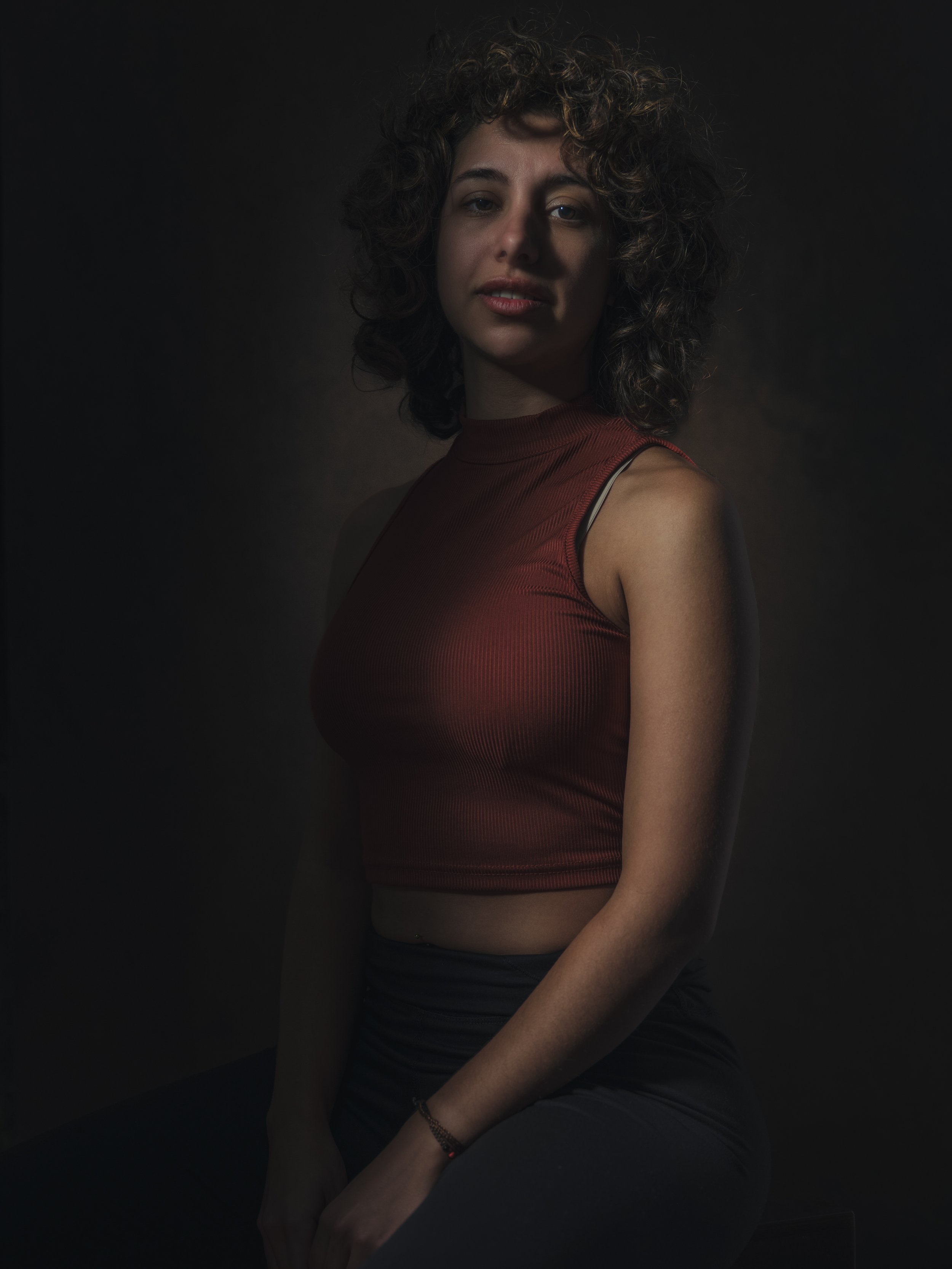 A woman with curly hair wearing a sleeveless red top and black pants standing against a dark background.
