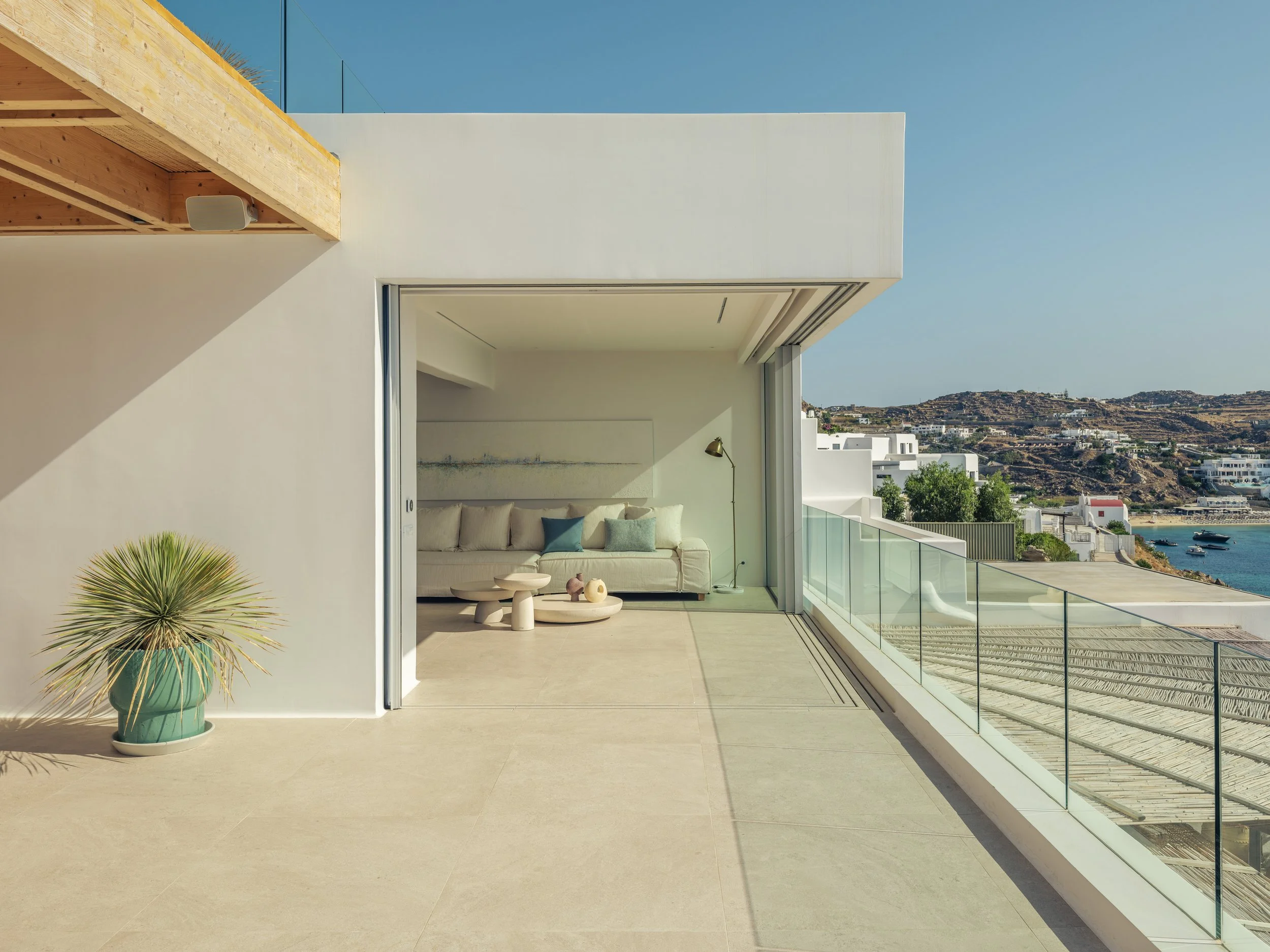Modern balcony with glass railing, outdoor seating, potted plant, and view of hills, buildings, and ocean.
