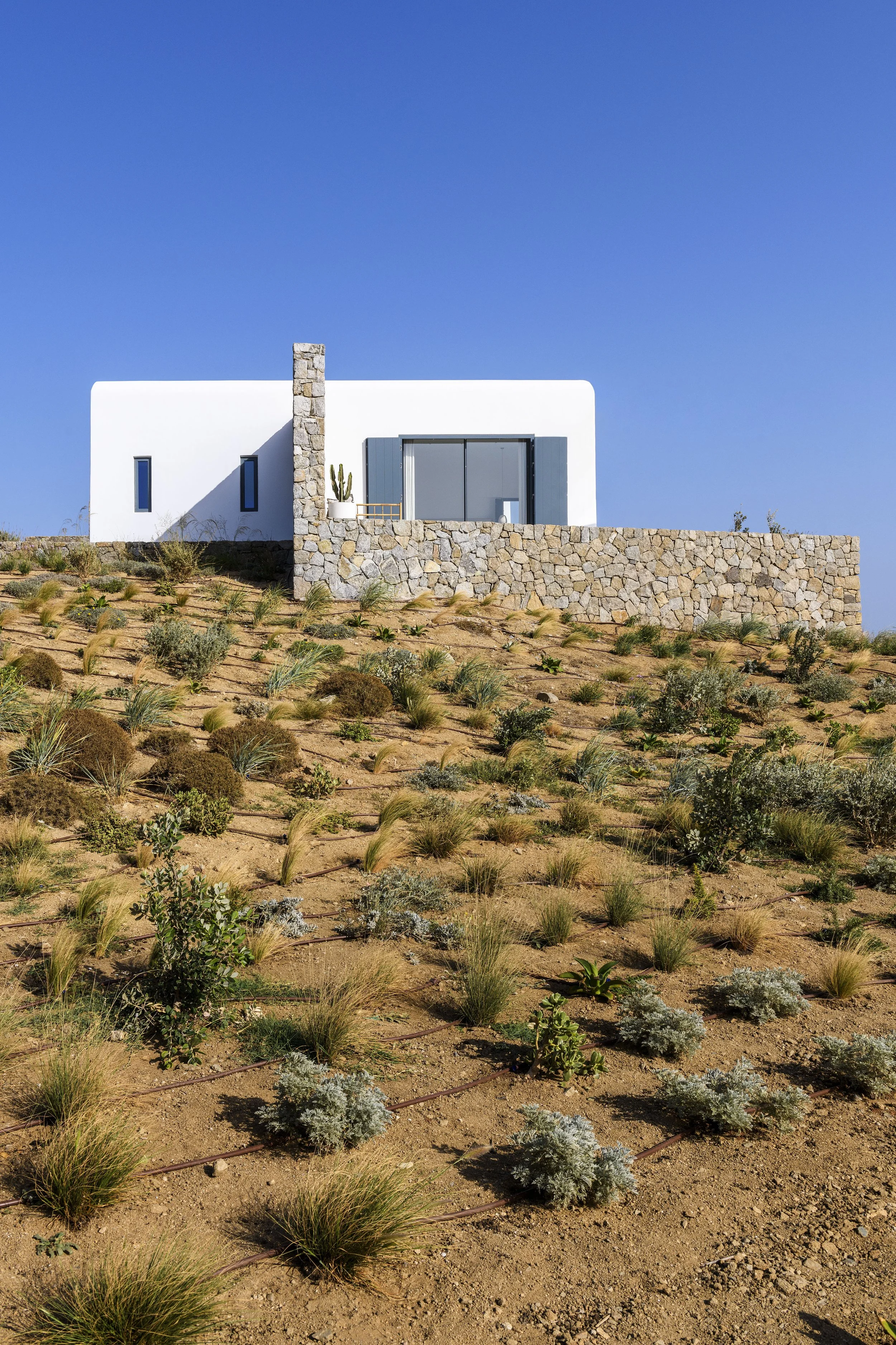 Modern white house with stone accents and a balcony, situated on a sloped terrain with desert vegetation and a clear blue sky.