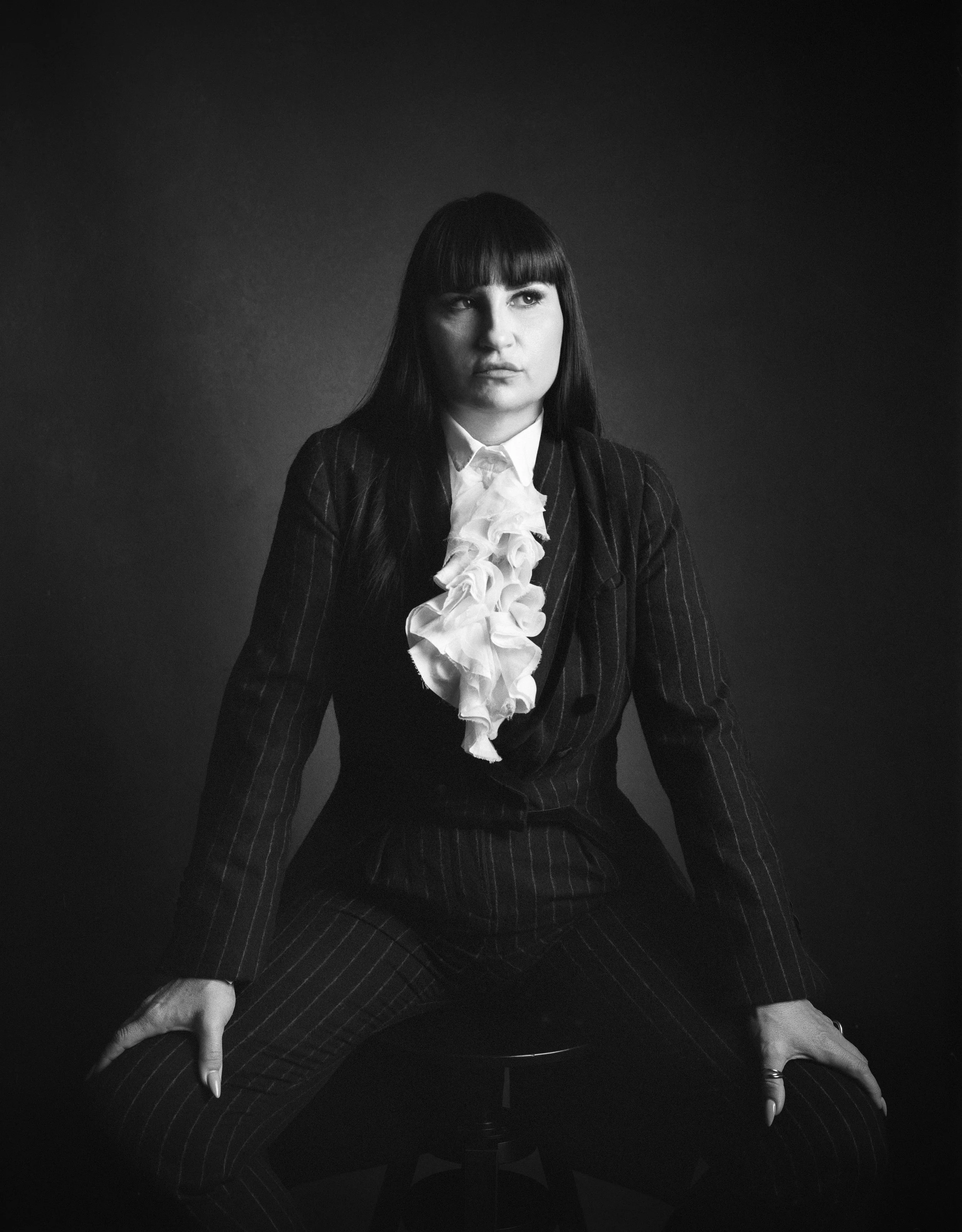 Black and white photo of a woman sitting with her legs apart, wearing a pinstripe suit with a ruffled blouse, looking to her right, against a dark background.