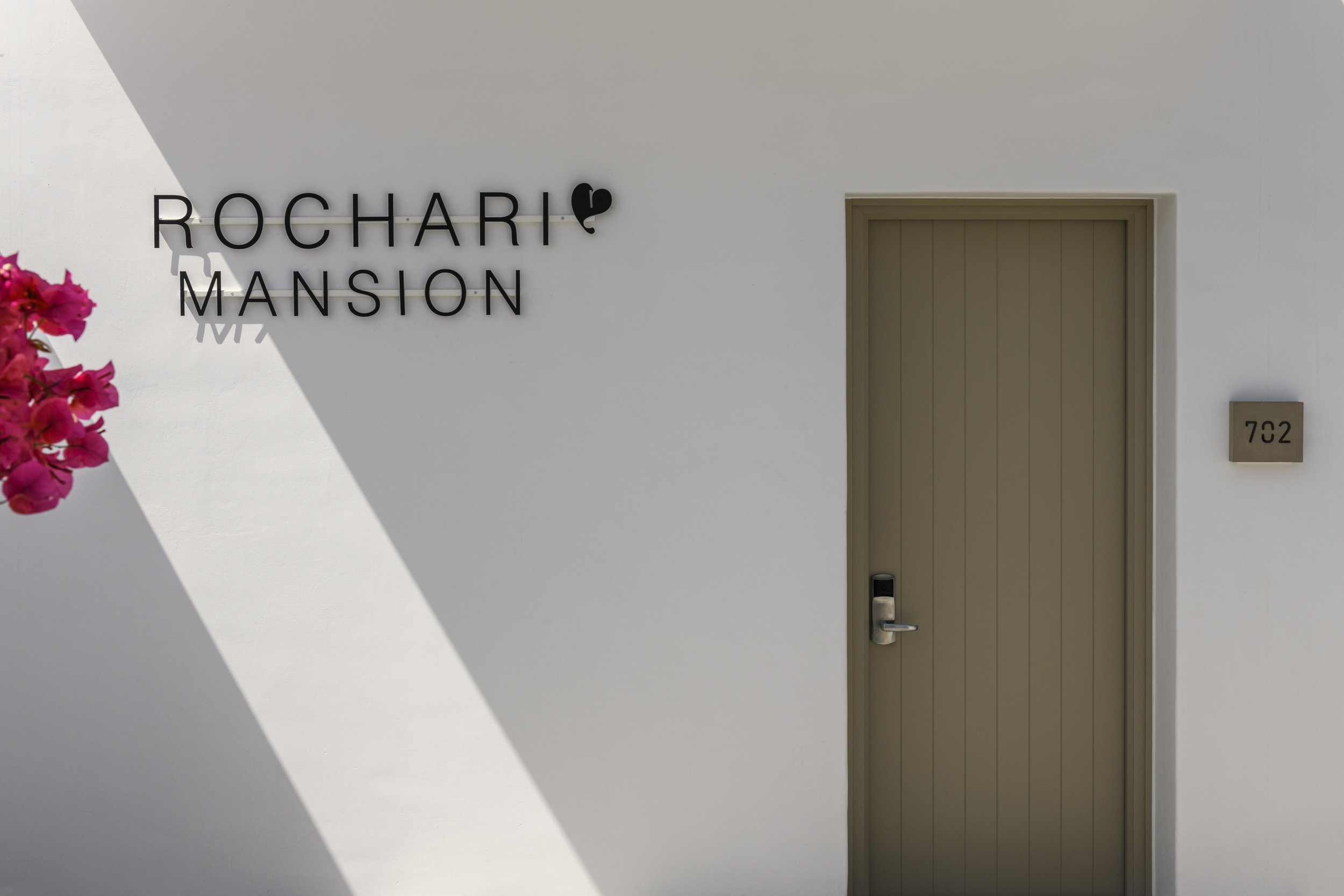 A white wall with black letters spelling 'ROCHARI MANSION' and a small black heart on the letter 'I'. A beige door with a door handle and lock is on the right side of the wall. Pink flowers are partially visible on the left side of the image.